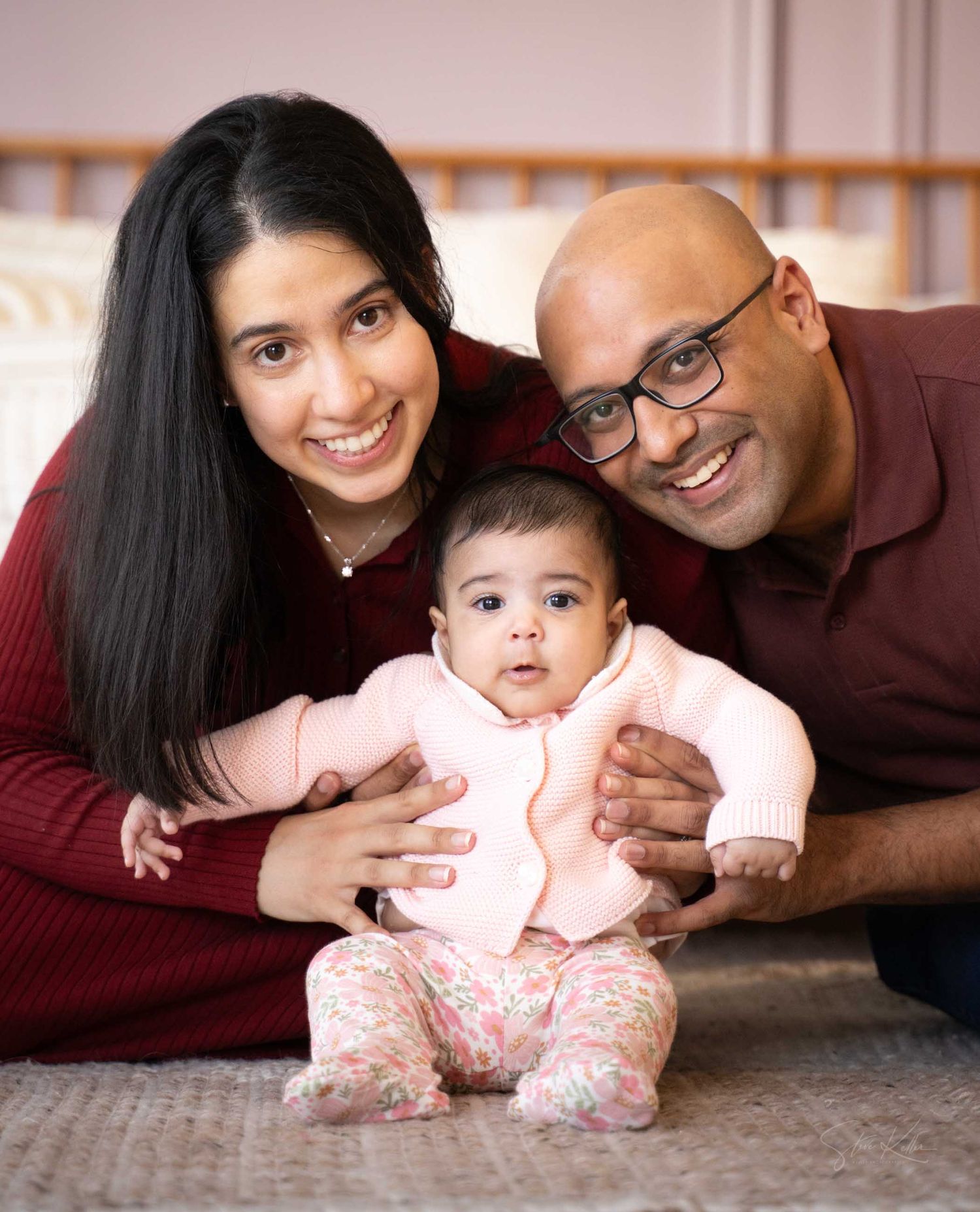 A smiling South Asian family with a mother, father wearing glasses, and their baby girl in pink sitting together on the floor.