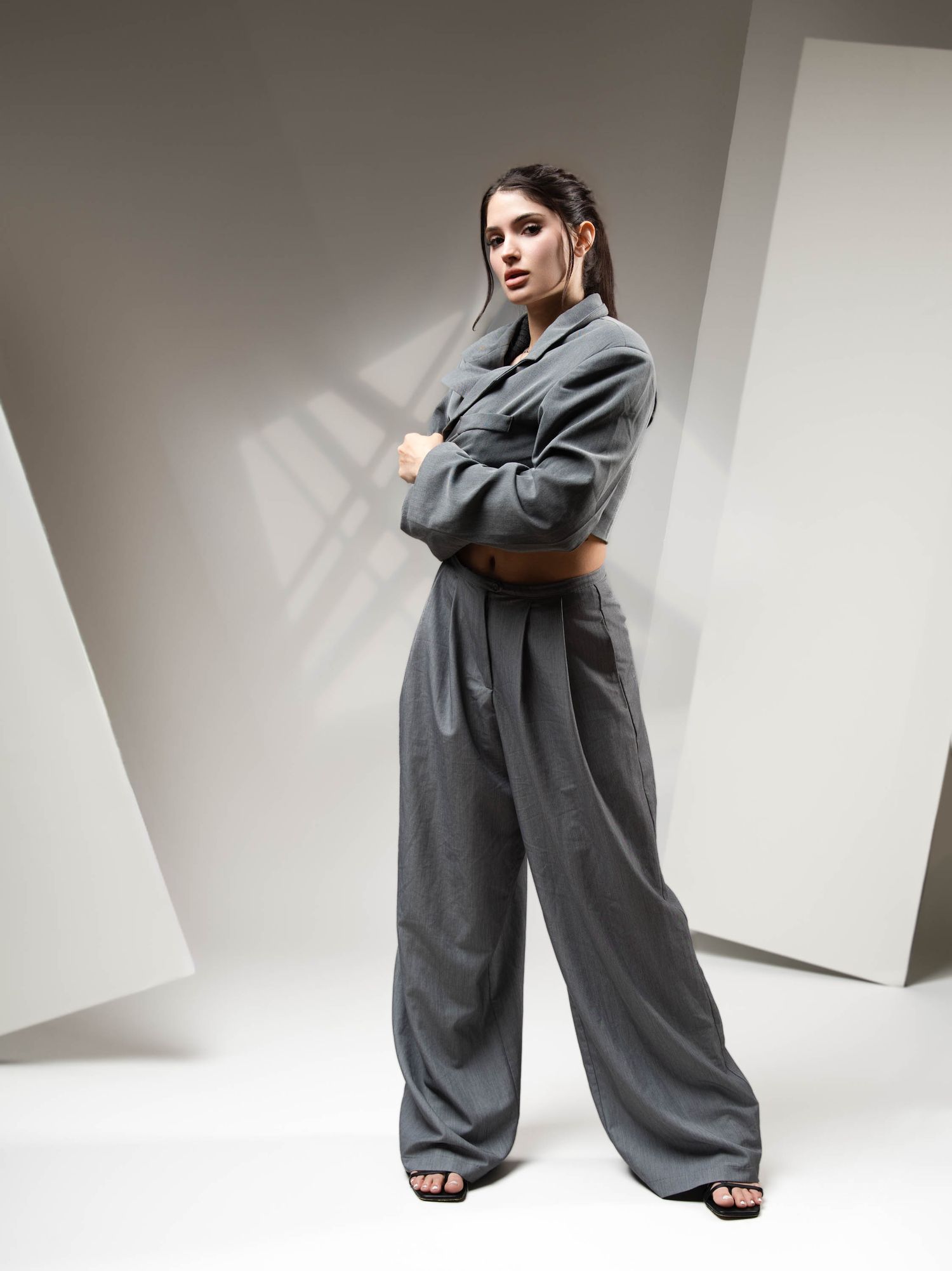 Fashion model in oversized gray blazer and wide-leg trousers poses against white studio backdrop with dramatic shadows.