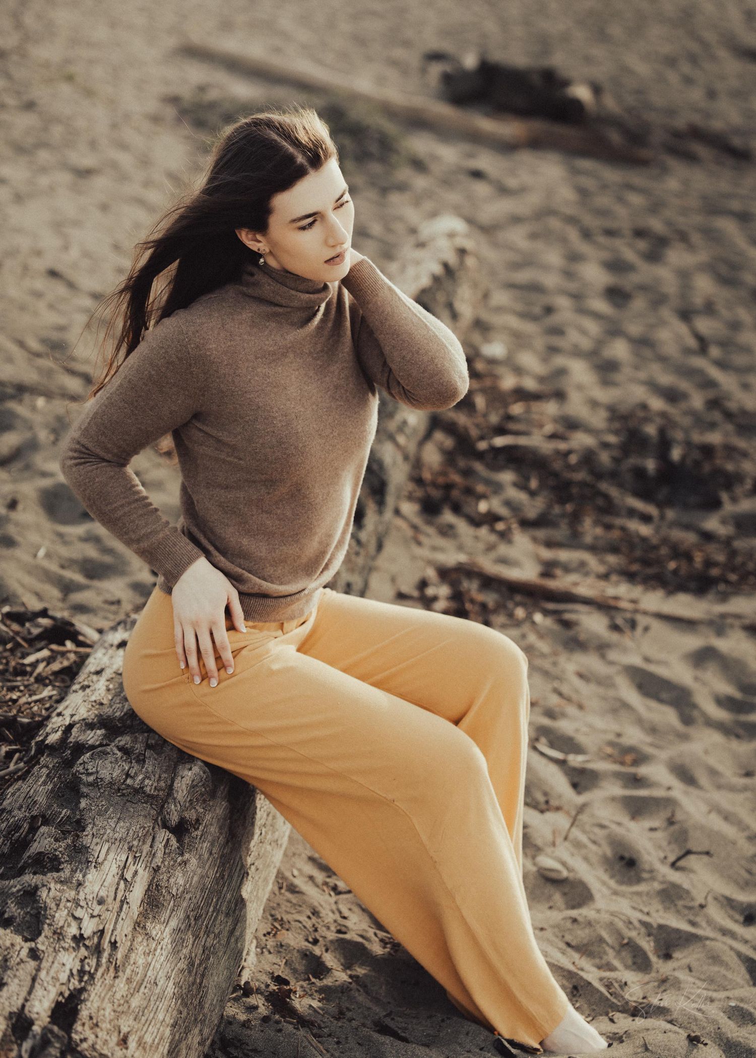 Figure wearing brown sweater and yellow pants sitting on driftwood at sandy beach during sunset.