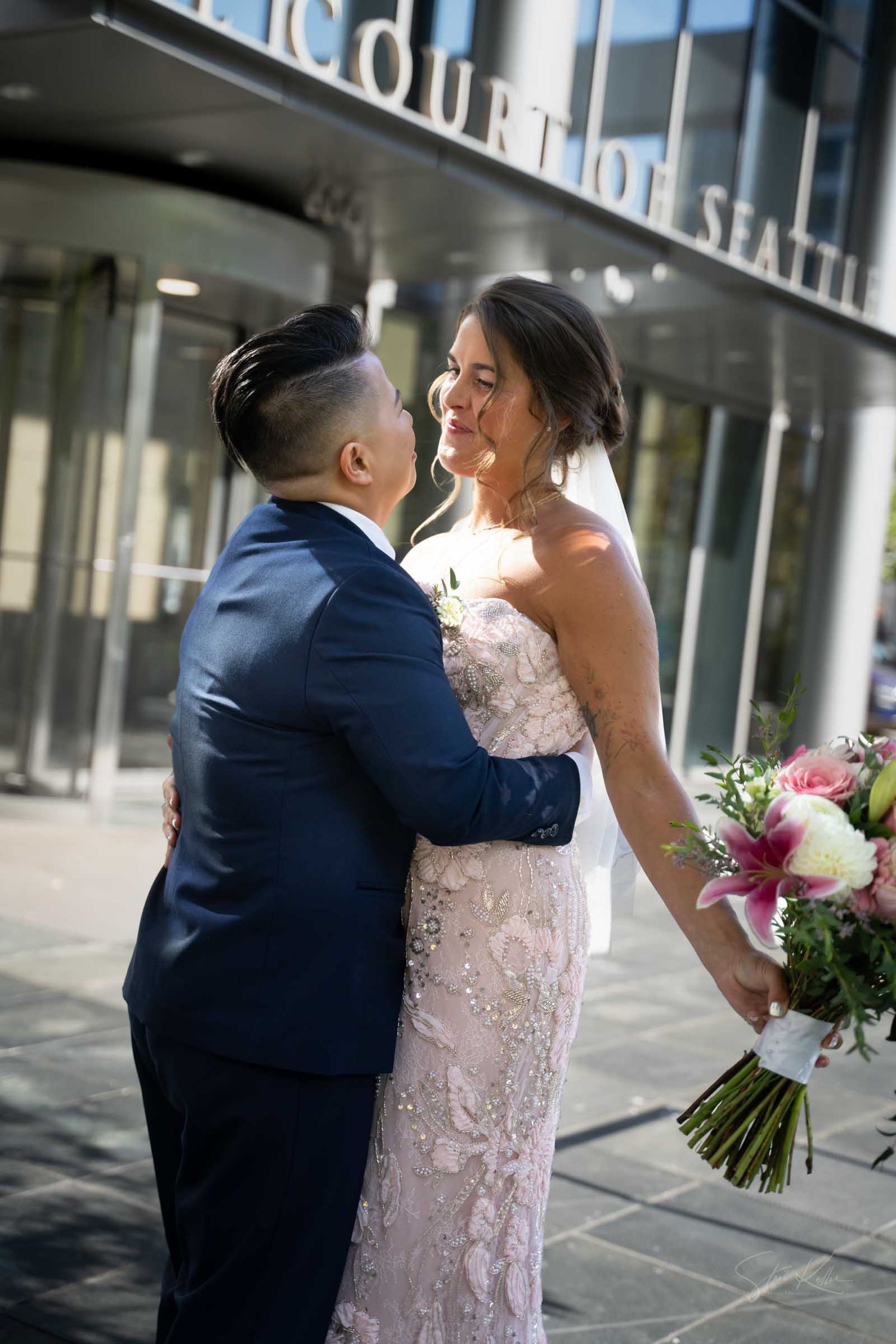 Newlywed couple embracing outside Seattle courthouse, bride in beaded gown holding pink bouquet, groom in navy suit.