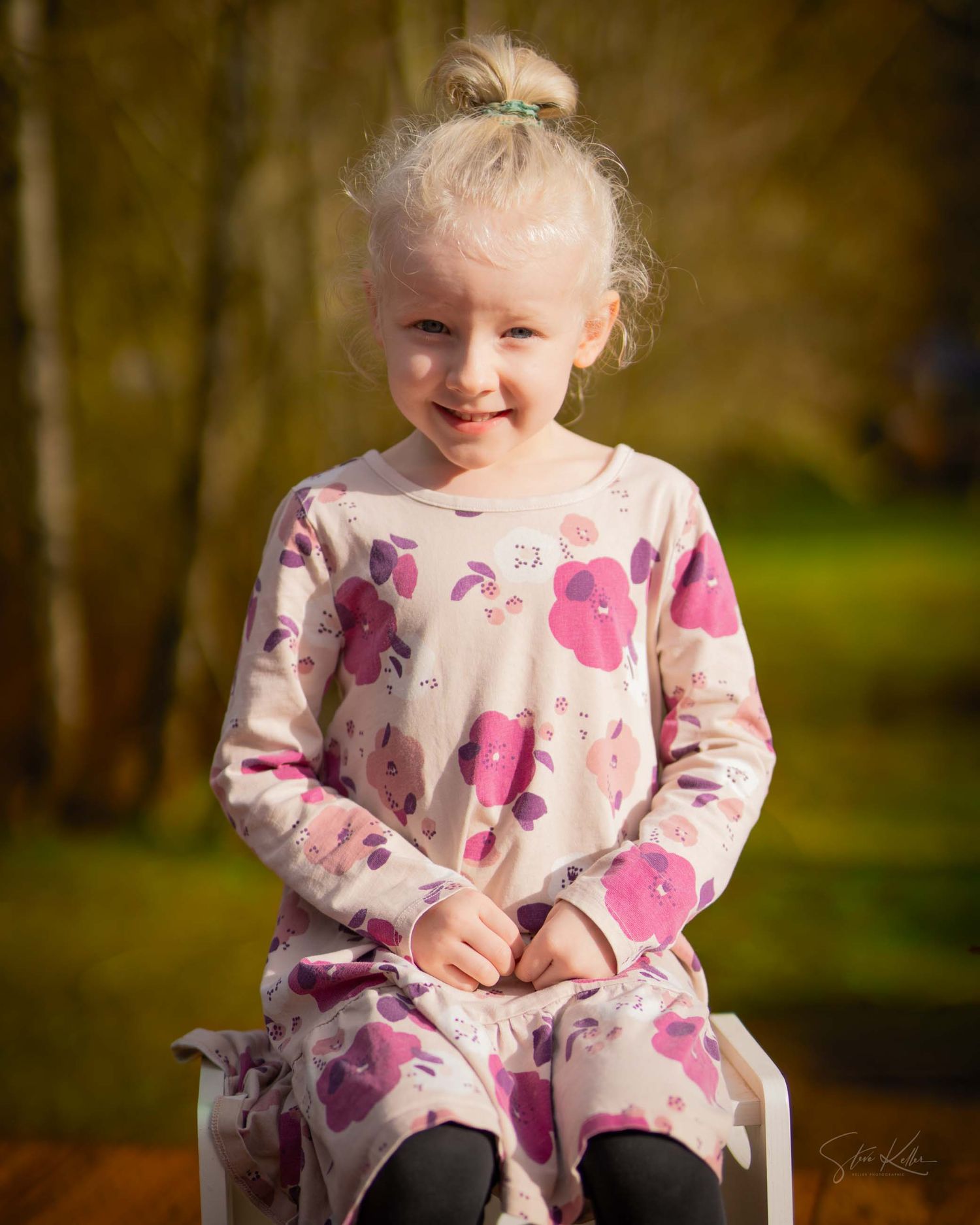 A child in a pink floral dress sits outdoors during golden hour lighting with a natural background.