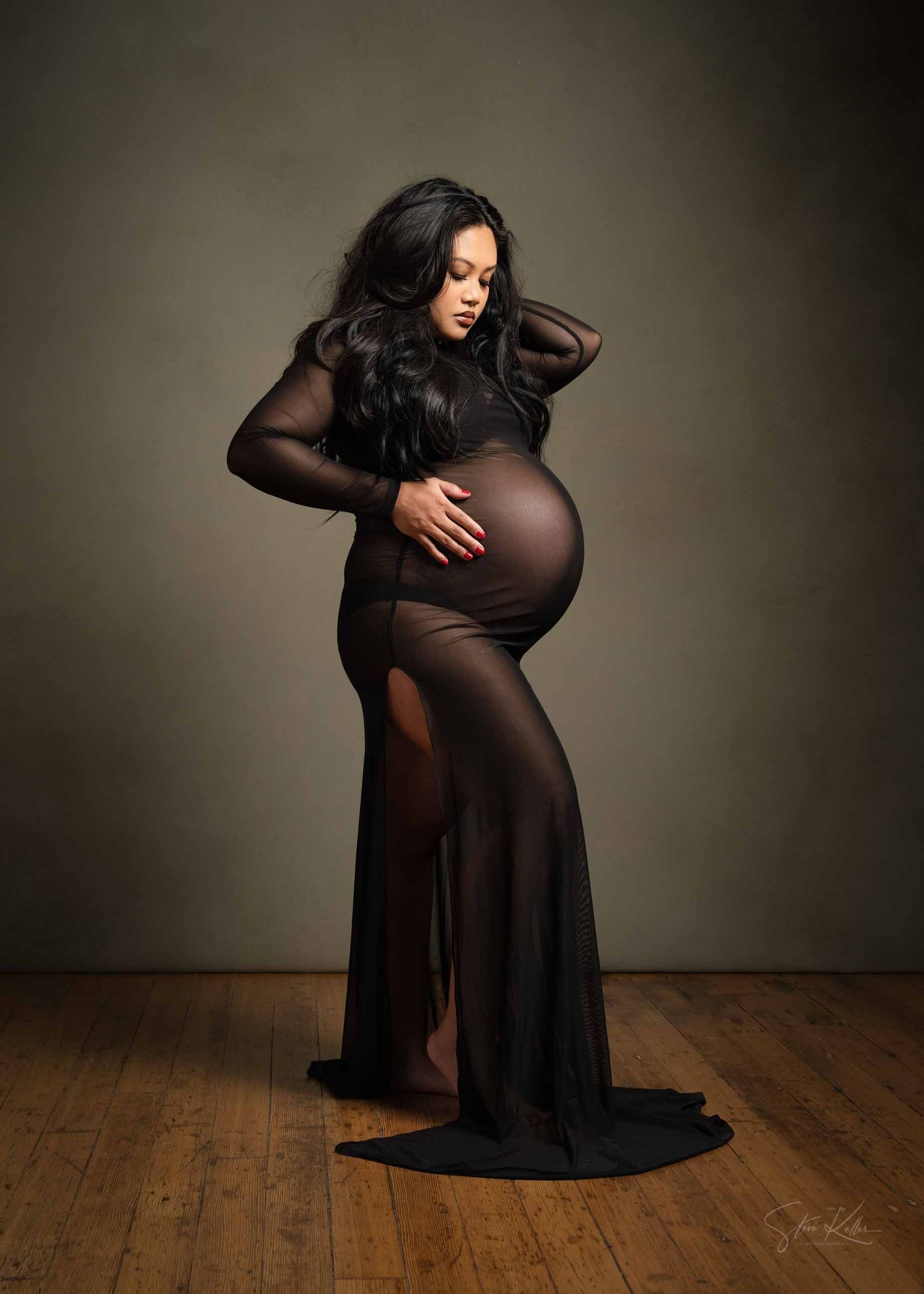 Artistic maternity portrait of an expectant mother in a flowing black sheer gown against a dark backdrop.