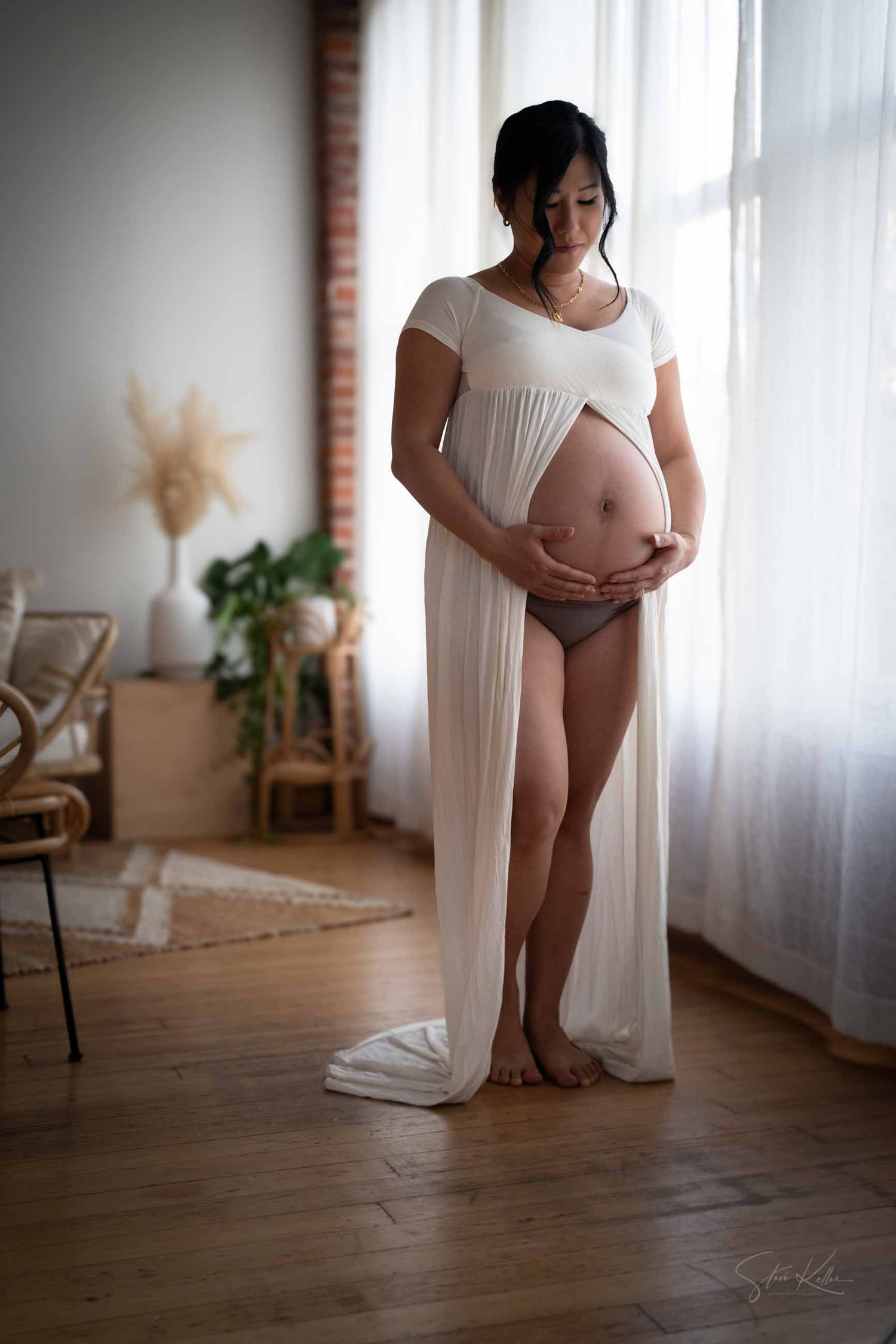 An expectant mother in a flowing white dress stands by a window in a minimalist room with natural decor.