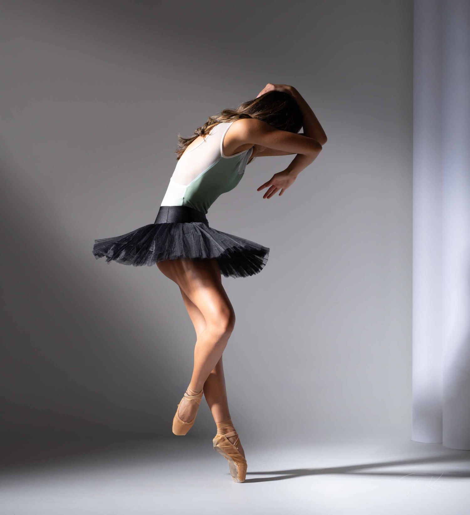 Ballet dancer in black tutu performs graceful movements in a minimalist studio setting with dramatic lighting.