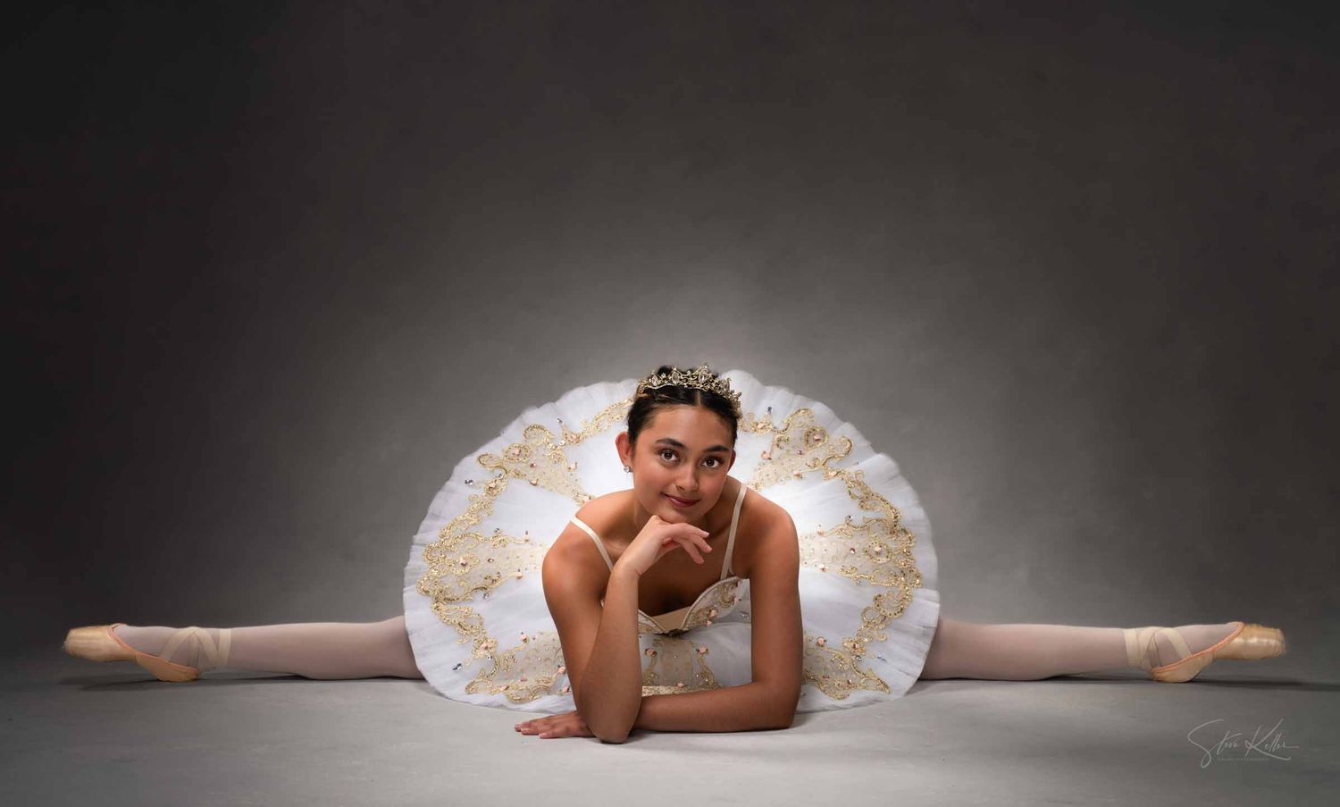 A ballet dancer performs a split on a dark stage with dramatic lighting and white tutu costume.