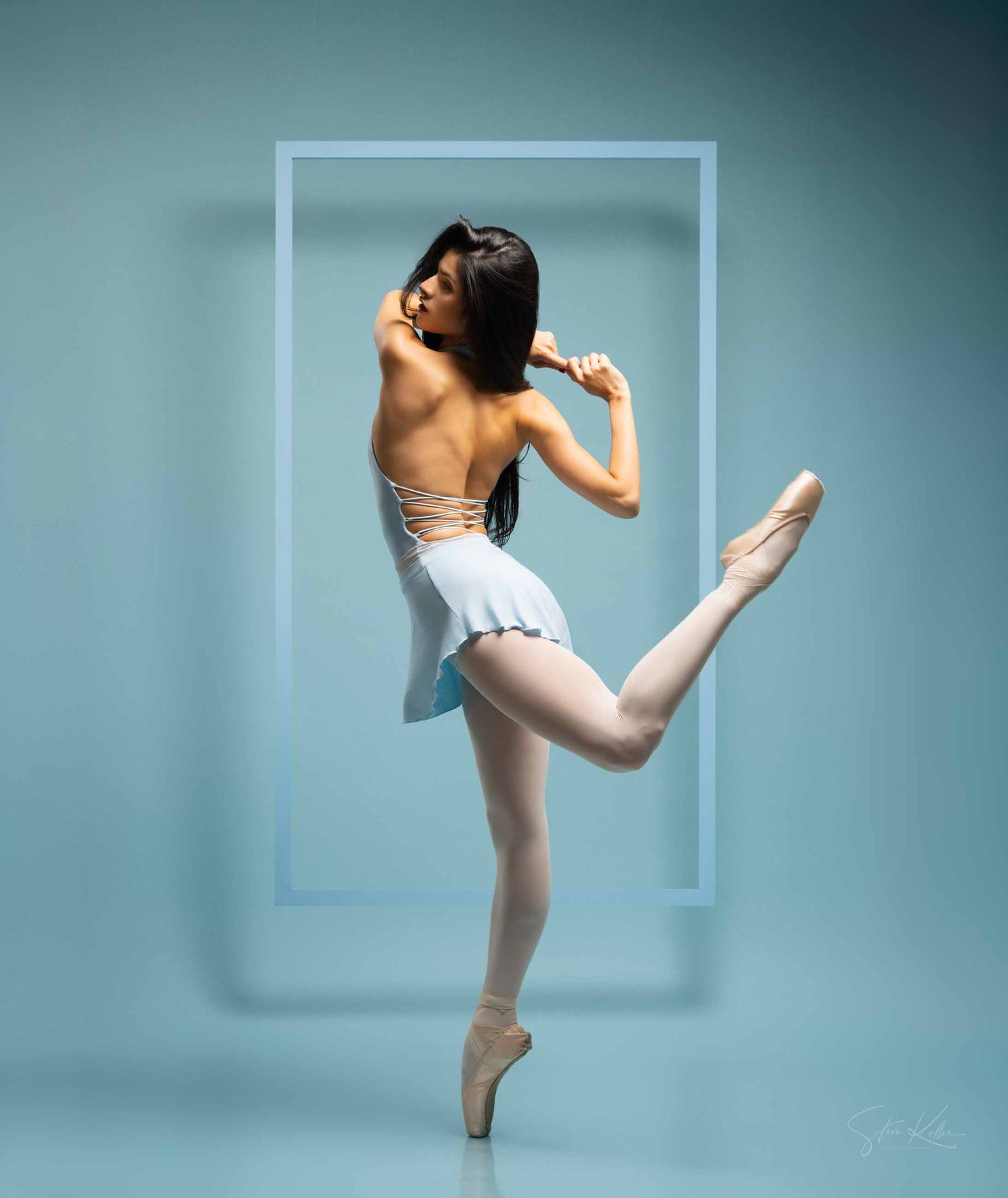 A ballet dancer in white performs a graceful high kick against a teal-colored studio backdrop.