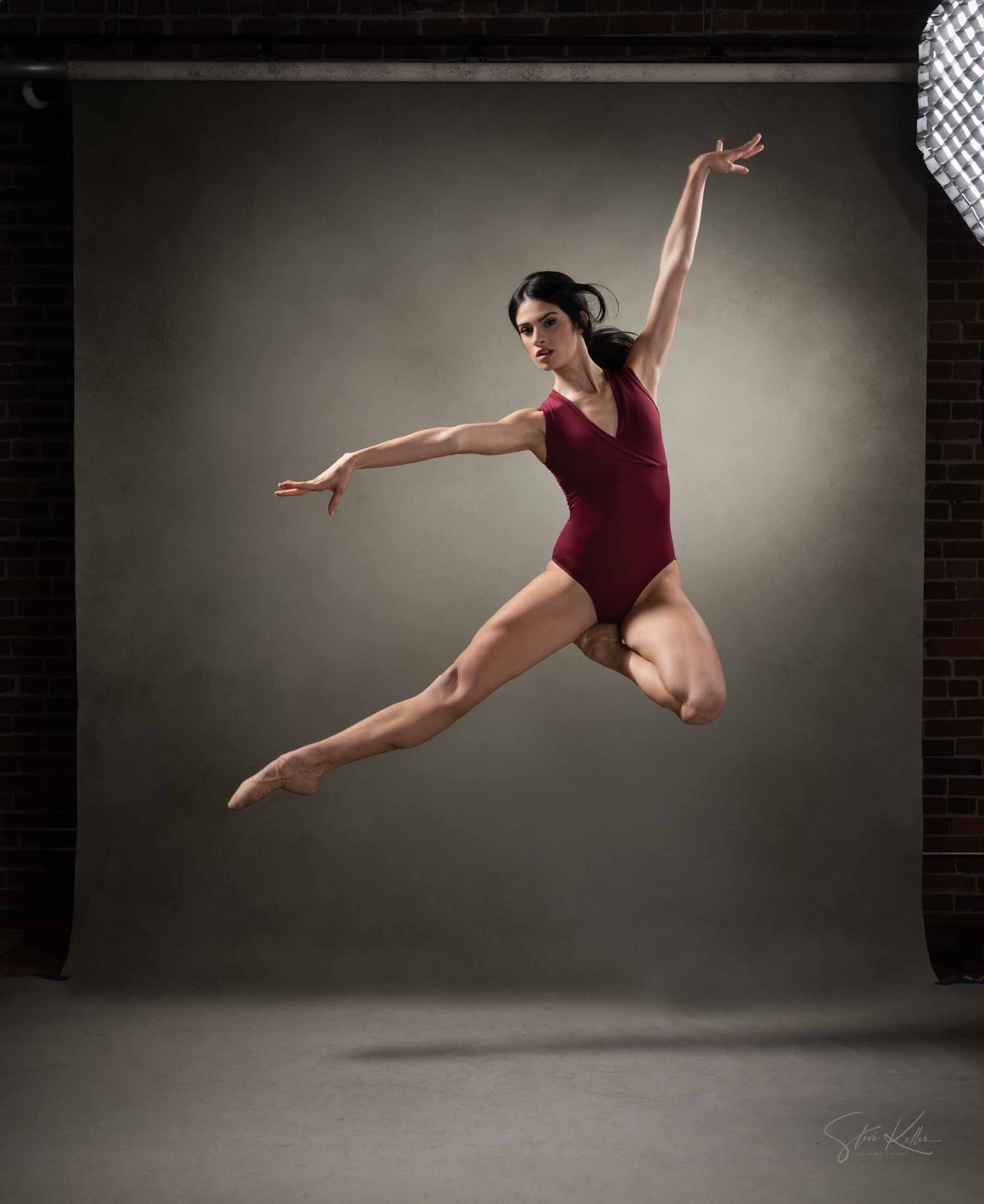 A dancer in burgundy leotard performs graceful leaps and poses in a dramatic studio setting against a dark background.