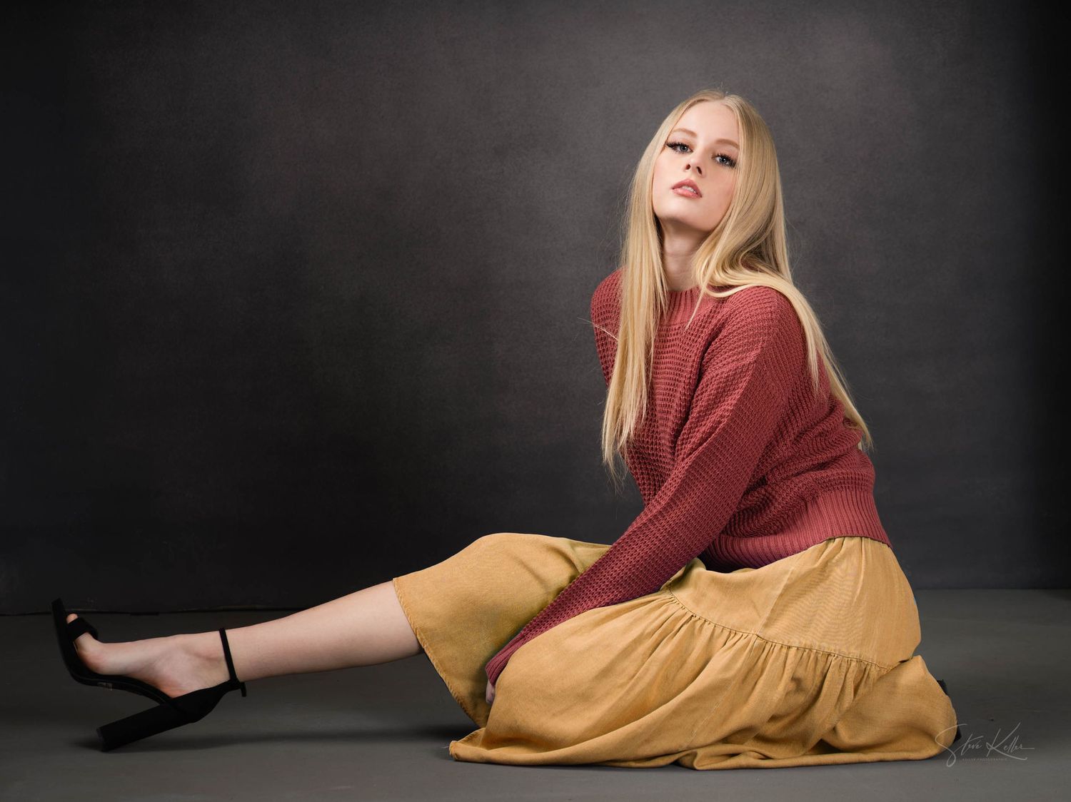 Young blonde woman in red top and golden skirt sitting gracefully on floor in professional studio setting.