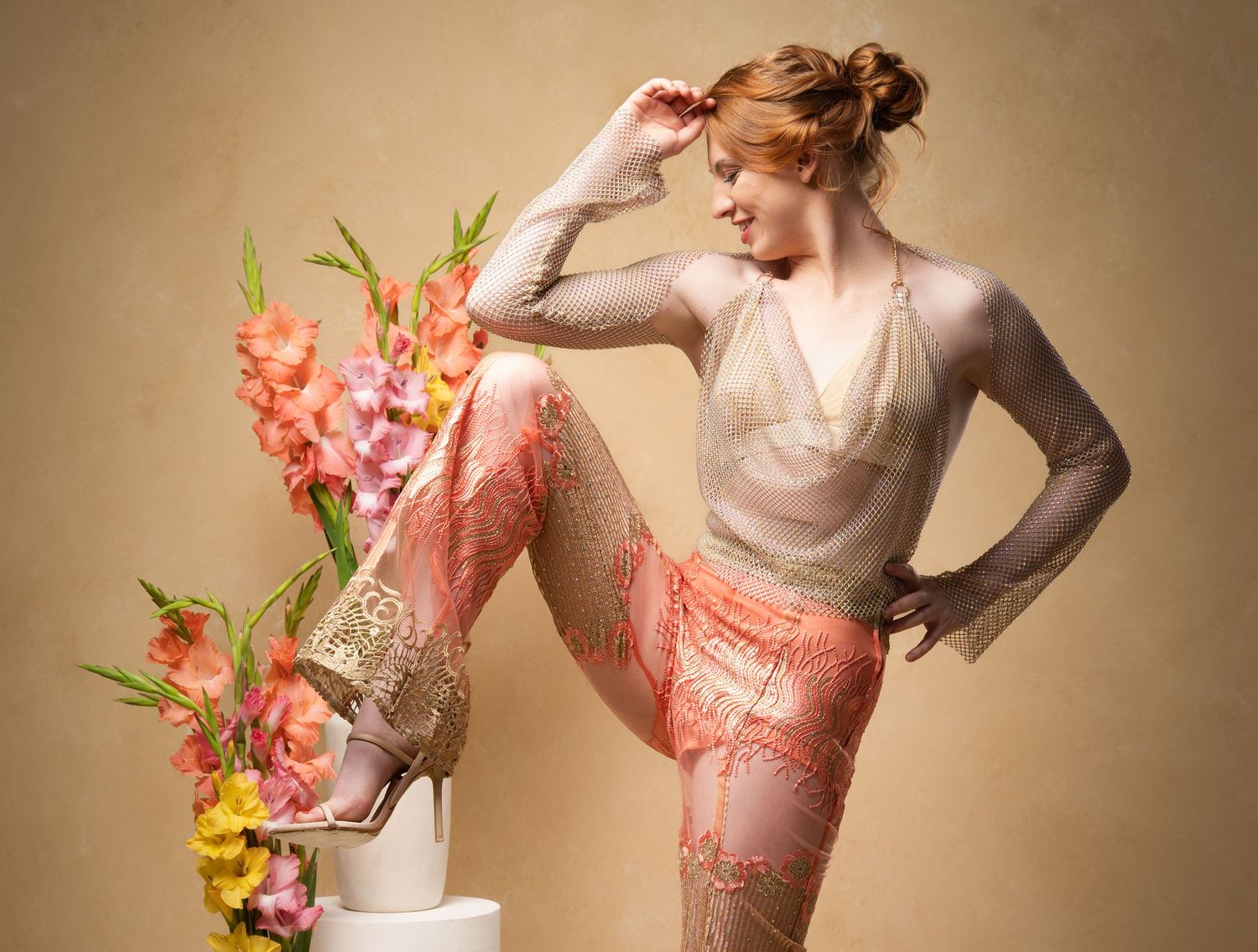 Model wearing ethereal floral applique gown in soft beige tones poses with delicate coral and yellow floral arrangements.