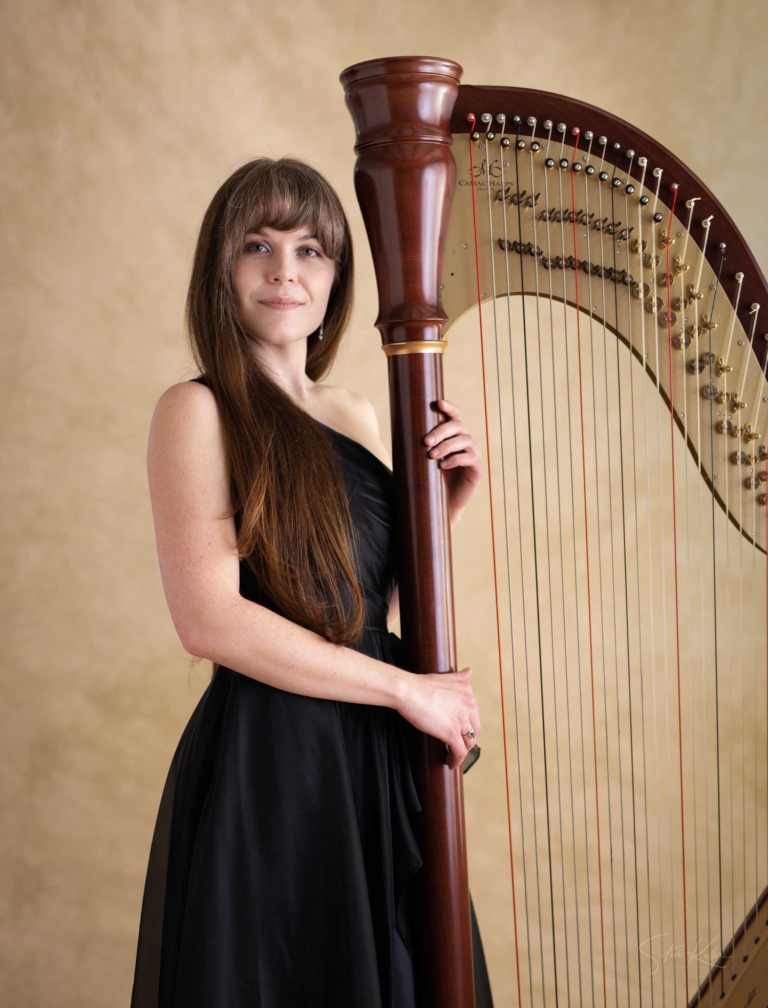 Female harpist in black dress standing beside a tall mahogany concert harp against a neutral backdrop.