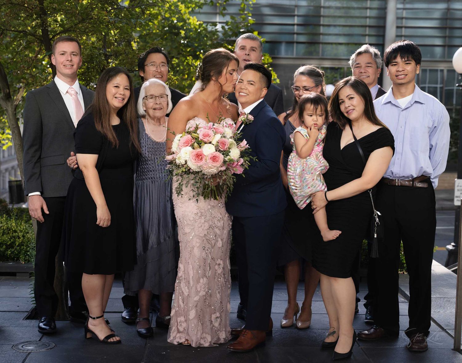 Wedding couple kissing surrounded by smiling family members outdoors at a urban venue.