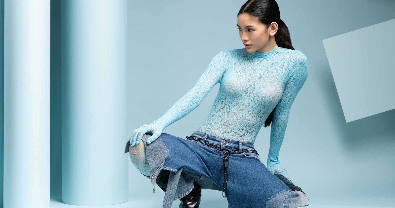 Model in a light blue mesh long-sleeve top and baggy jeans crouches against a pale blue studio backdrop.