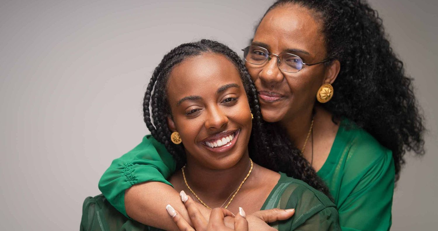 Smiling woman with braids and gold jewelry is embraced from behind by an older woman with glasses, both wearing green in a warm portrait.