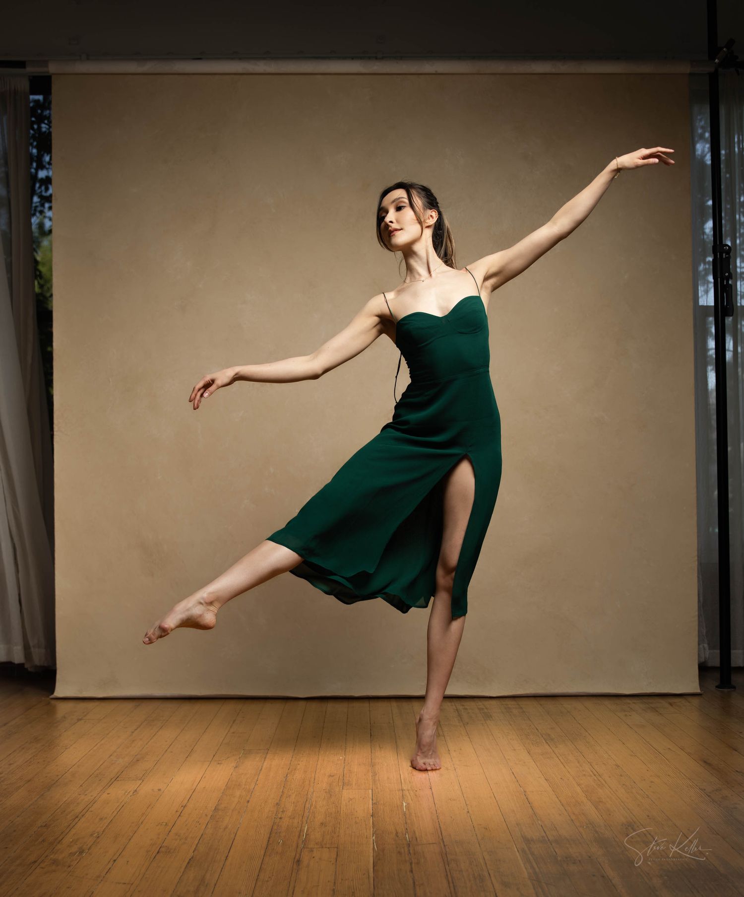 A ballet dancer in an emerald green dress performs an elegant pose on a wooden floor against a neutral backdrop.