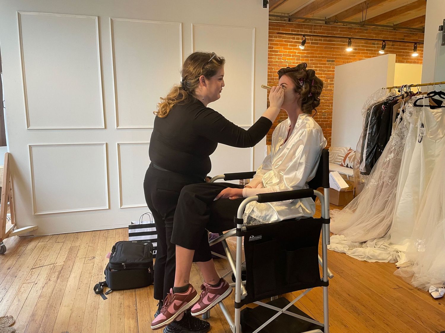 Makeup artist applying bridal makeup to a woman in a white robe near wedding gowns in a bridal studio.