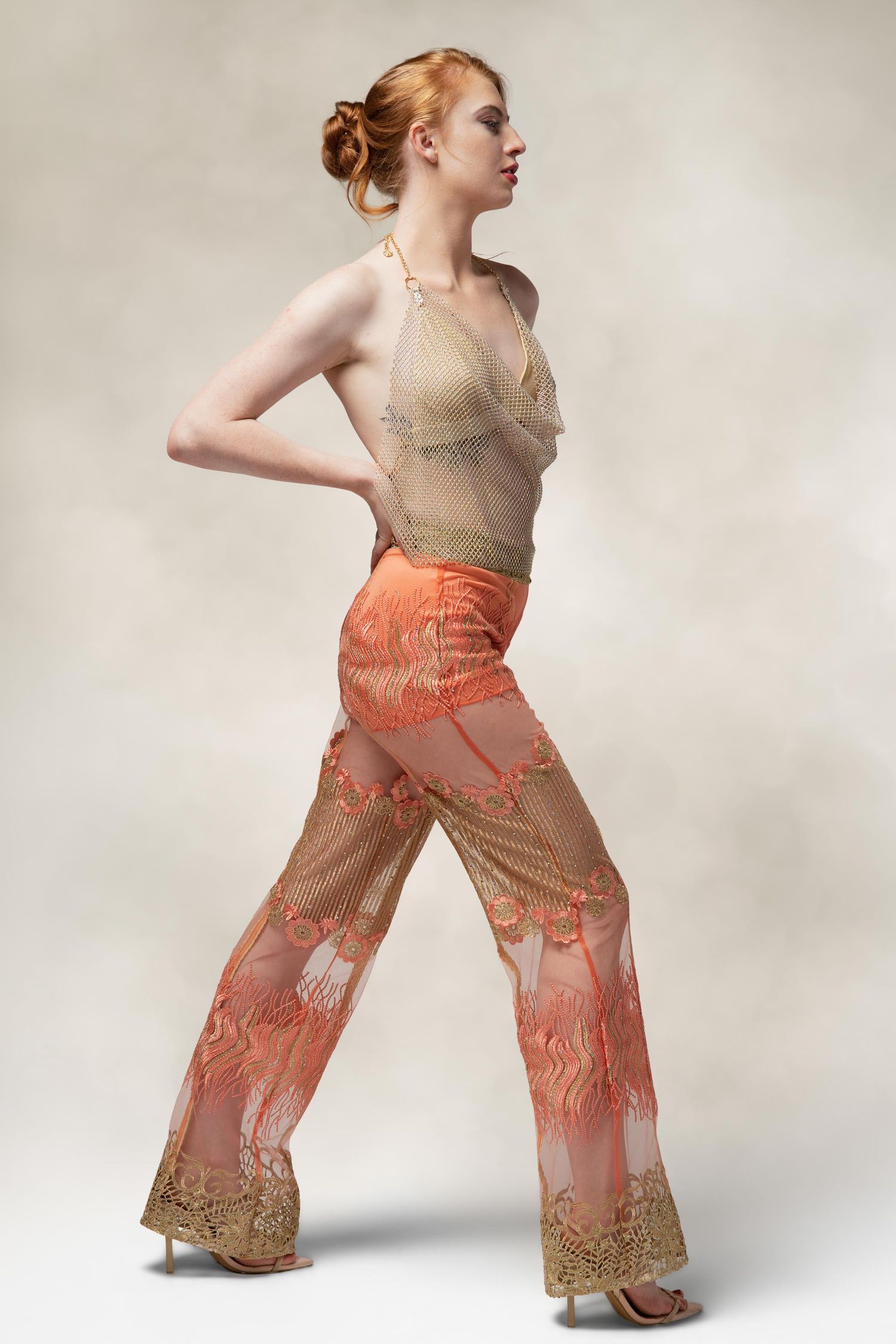 Fashion model poses in sheer orange patterned palazzo pants and beige top against neutral studio background.