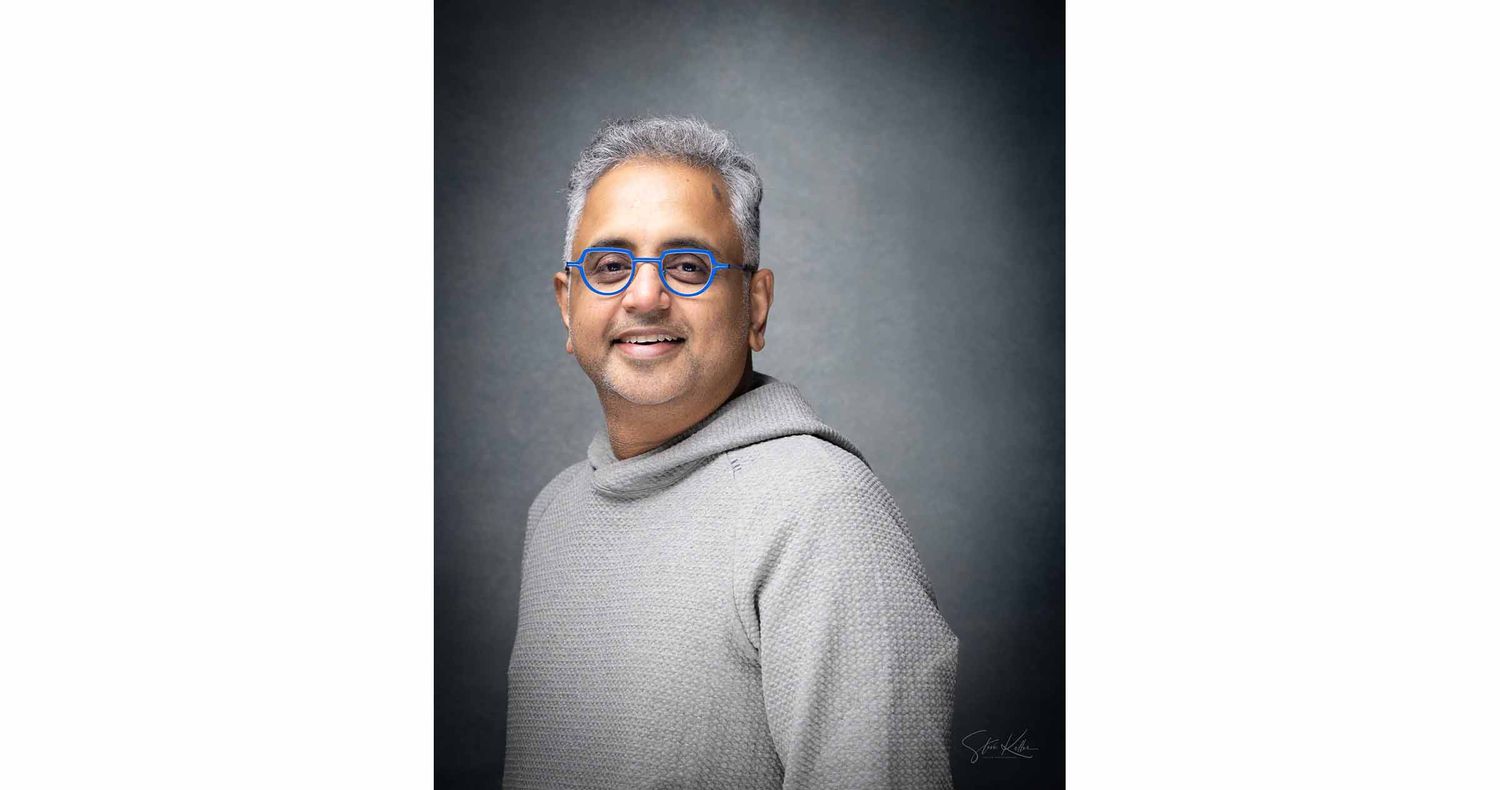 Smiling man with gray hair and blue-tinted round glasses wears a light gray hoodie in a studio portrait against a textured gray backdrop.