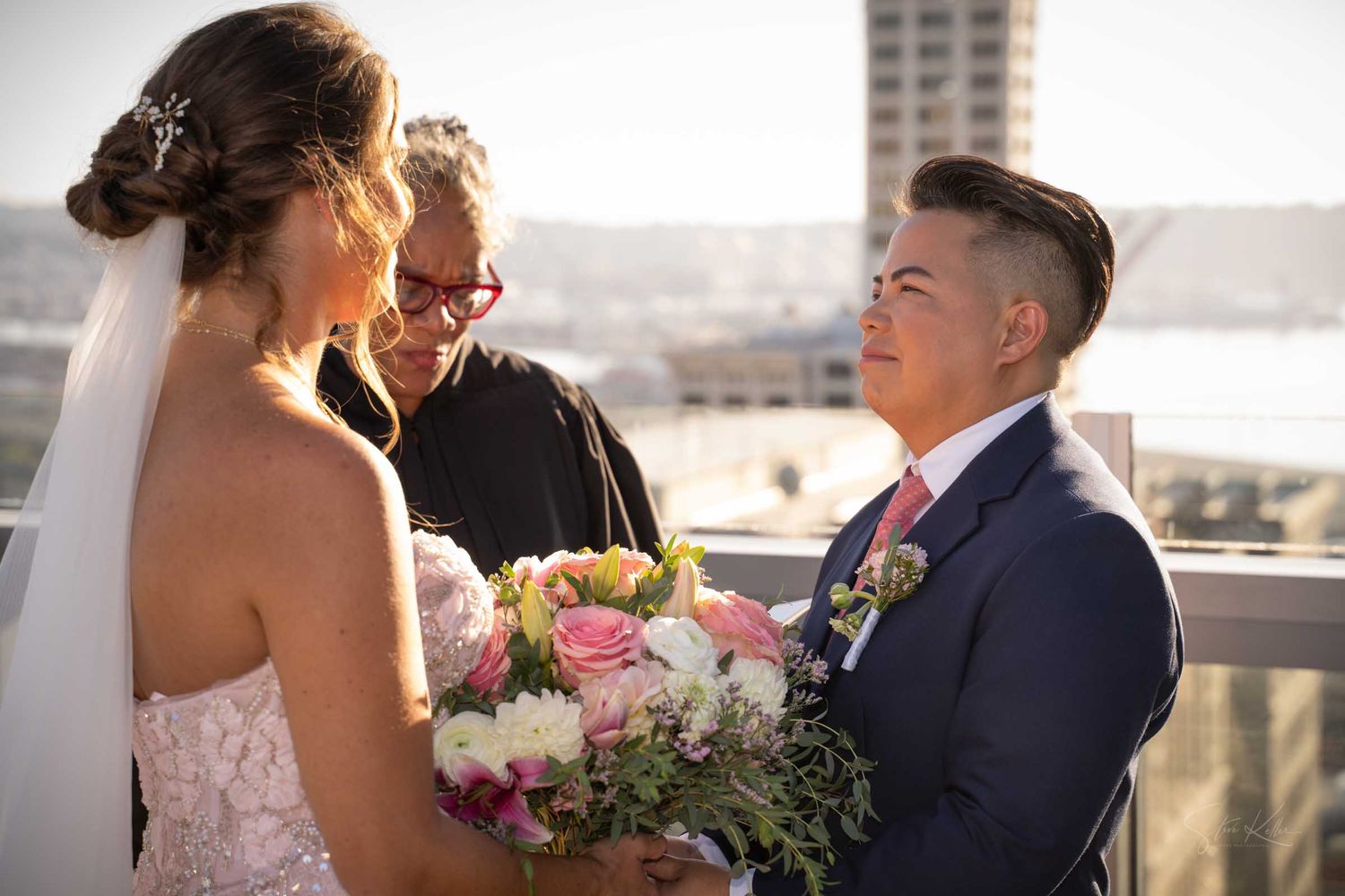 Two brides exchange vows on a rooftop with an officiant, one holding a pink and white wedding bouquet.