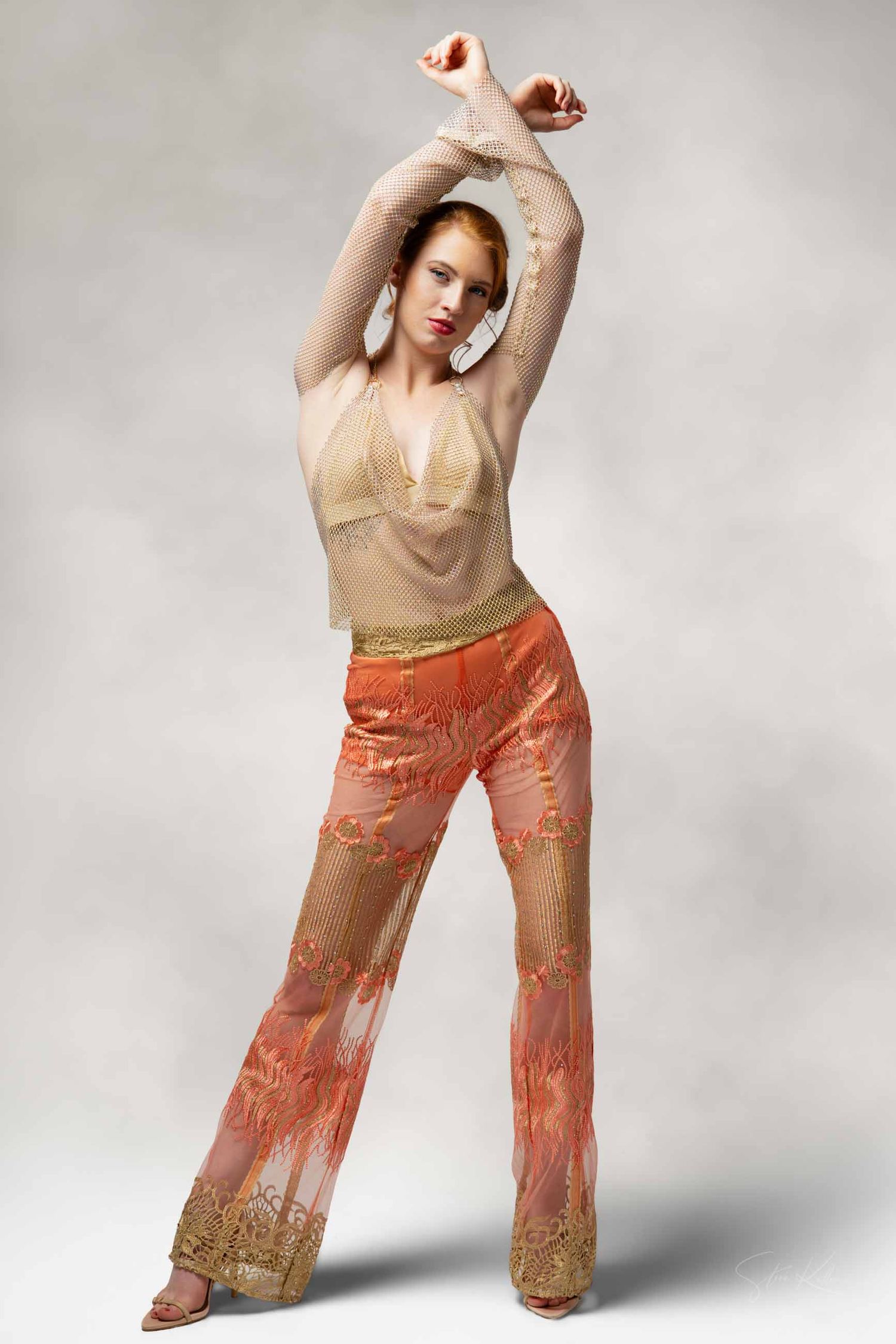 Model poses with raised arms in beige mesh top and coral patterned pants against neutral backdrop.