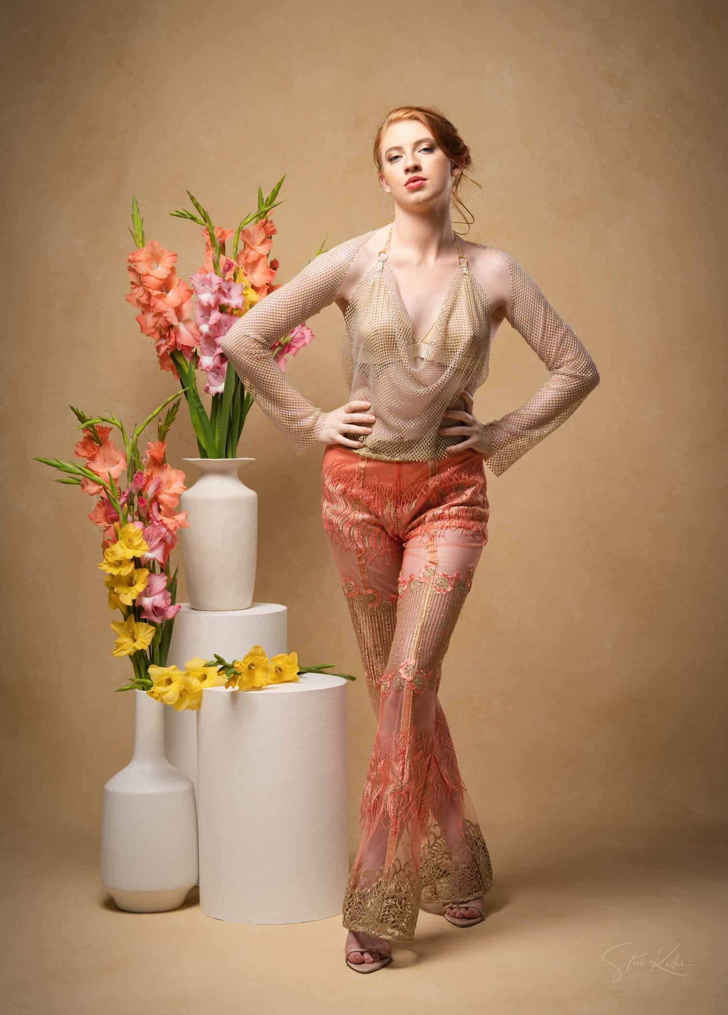 Fashion portrait with coral gladiolus flowers in white vases and model wearing beige top and shimmering coral pants.