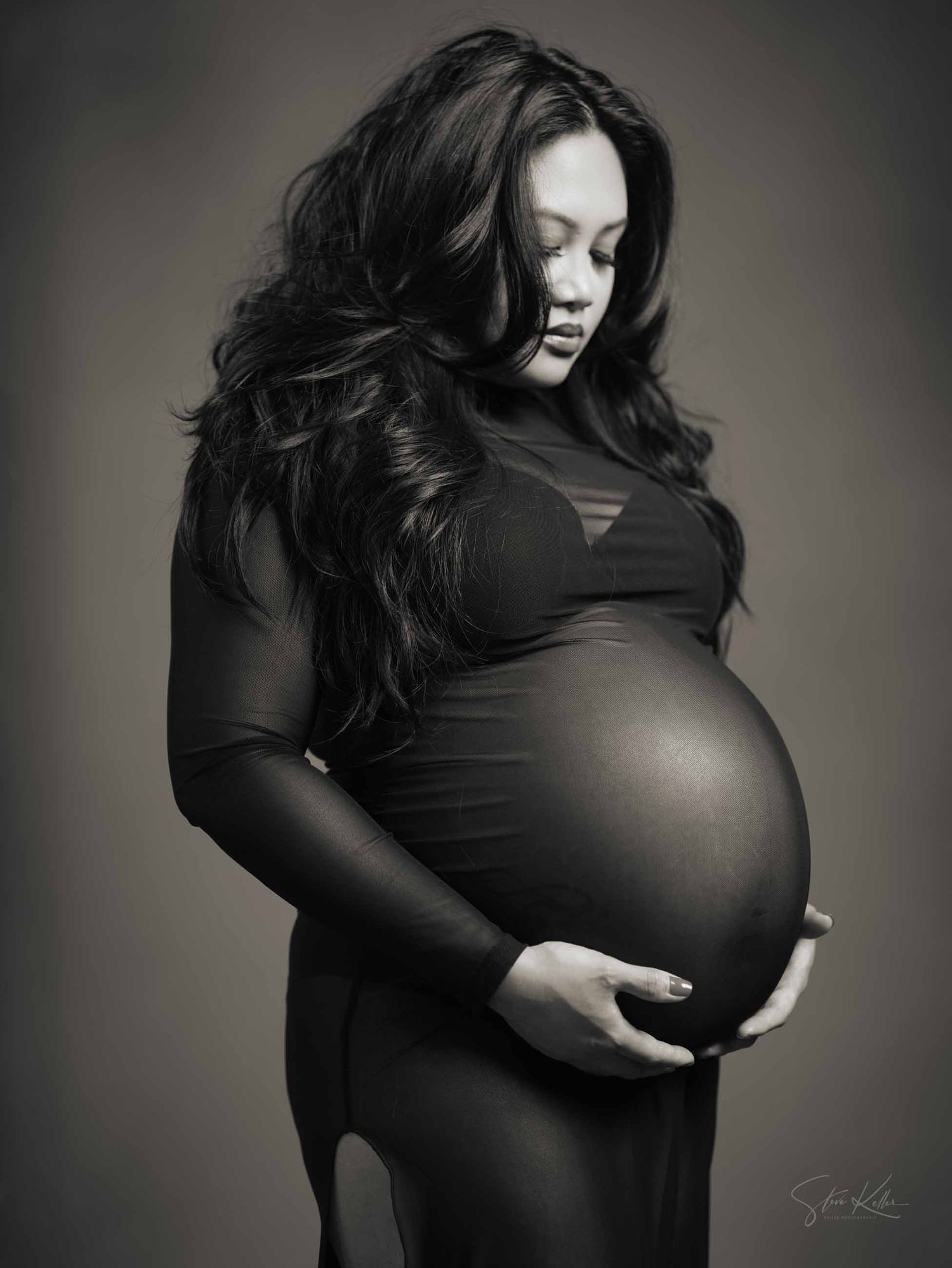 Artistic black and white maternity portrait of an expectant mother in a dark dress against a shadowy background.