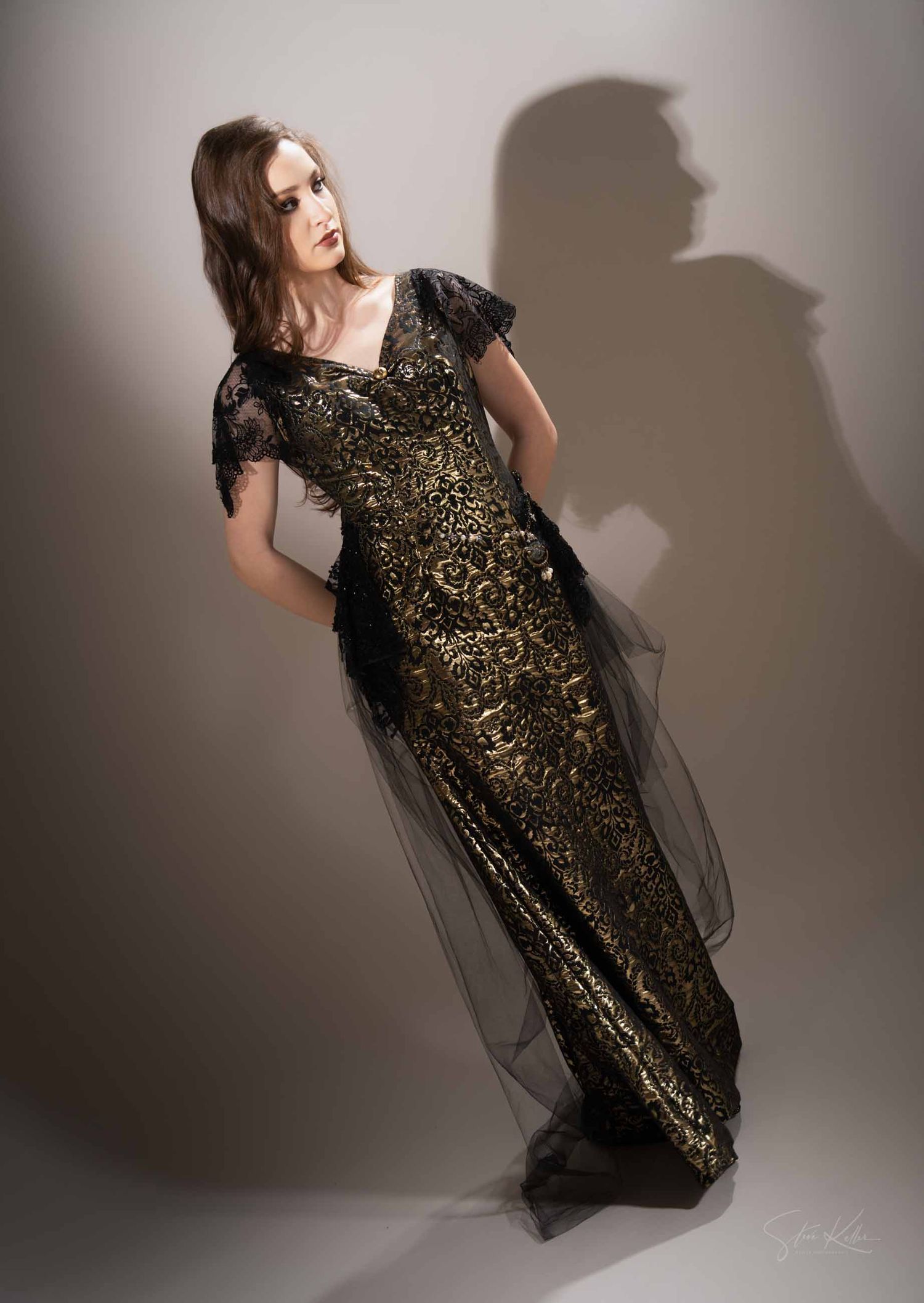 A model poses in a black and gold sequined dress with dramatic tulle details against a gray studio backdrop.