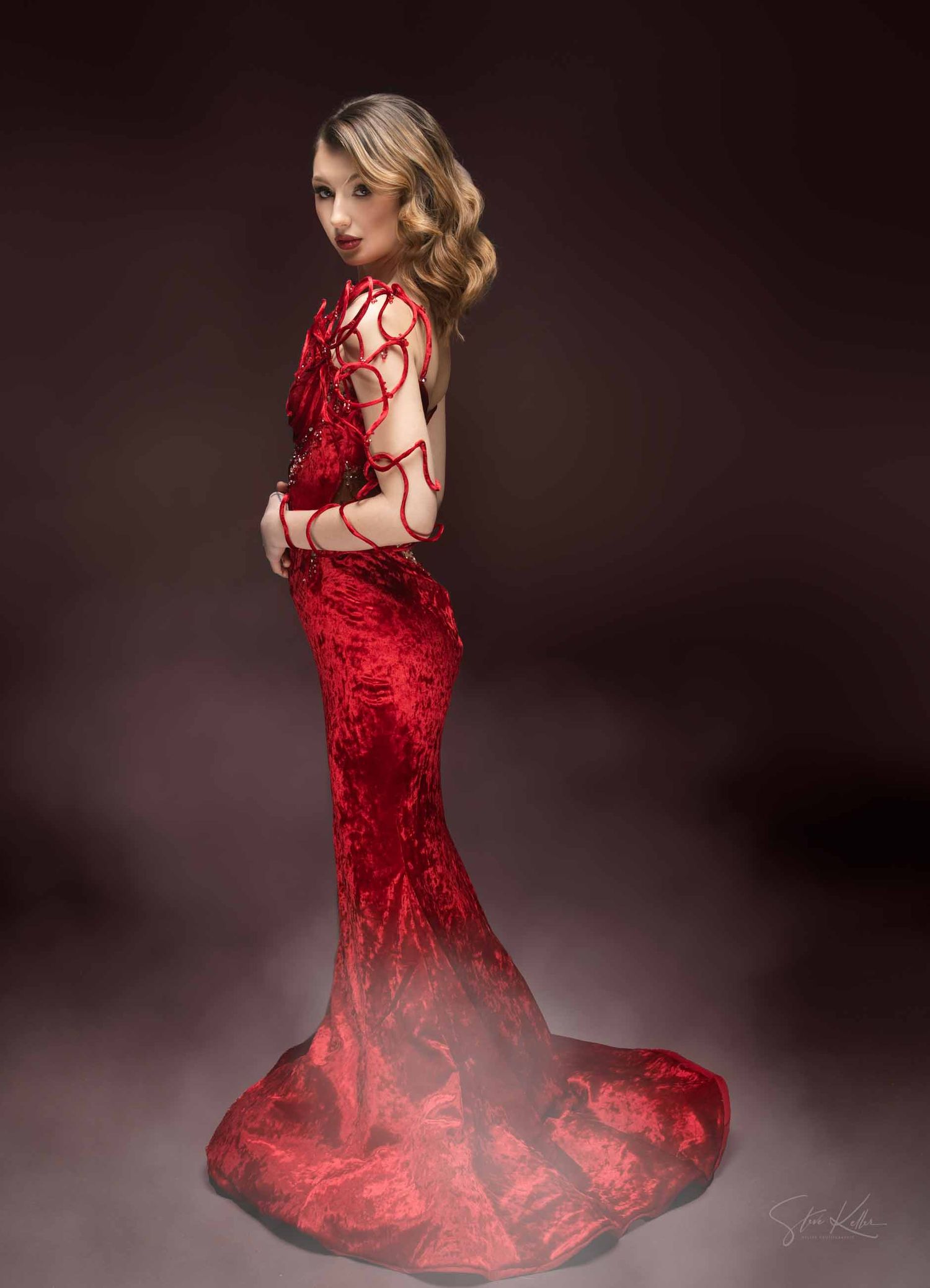 A model in a stunning red lace evening gown poses dramatically against a dark background in a fashion photoshoot series.
