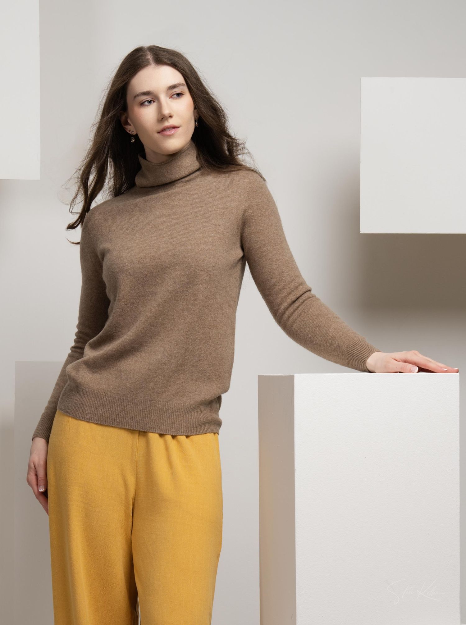 Woman in camel turtleneck sweater and yellow pants leaning against white geometric backdrop.