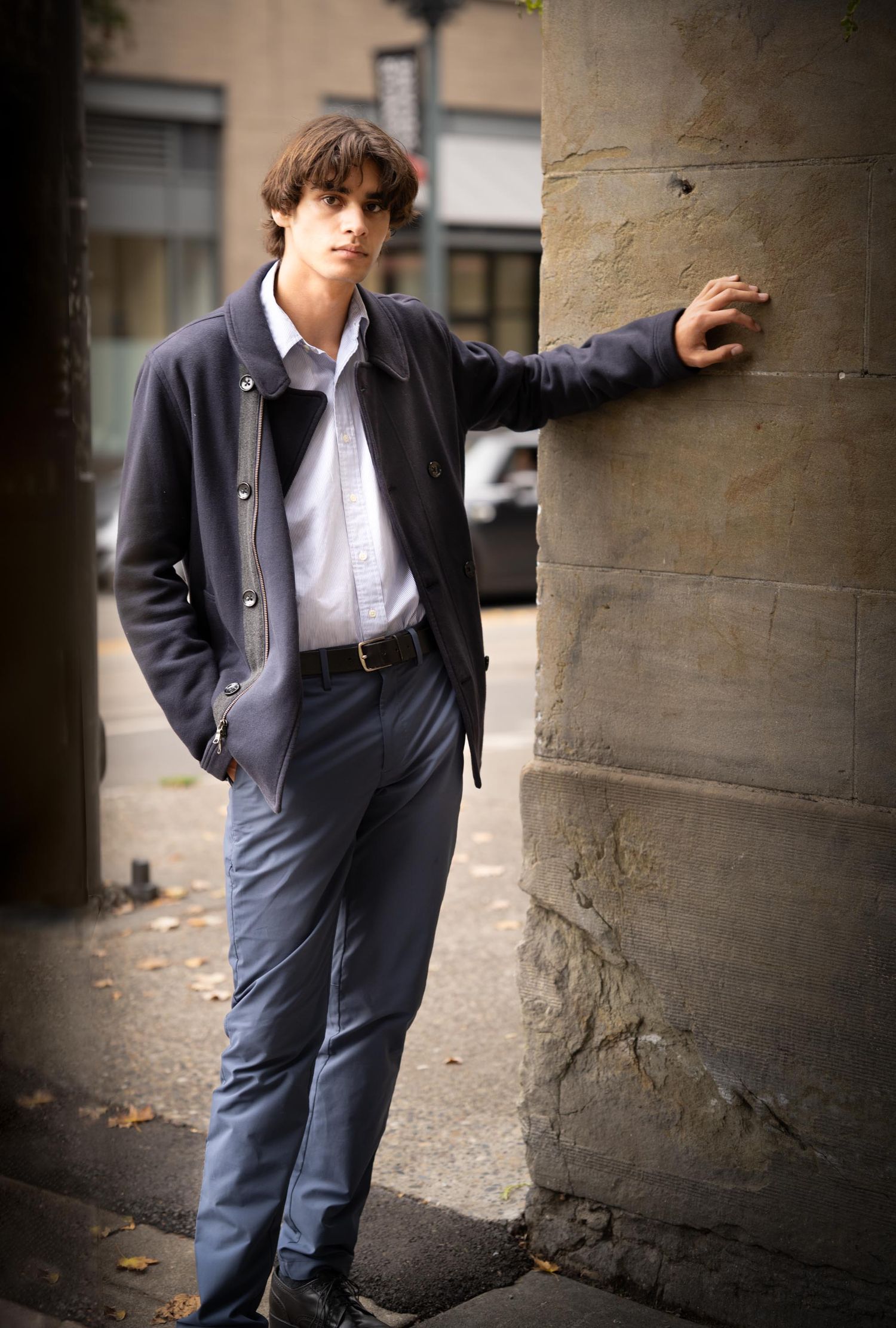 A person in a navy peacoat and dress pants leans against a concrete pillar on an autumn day.