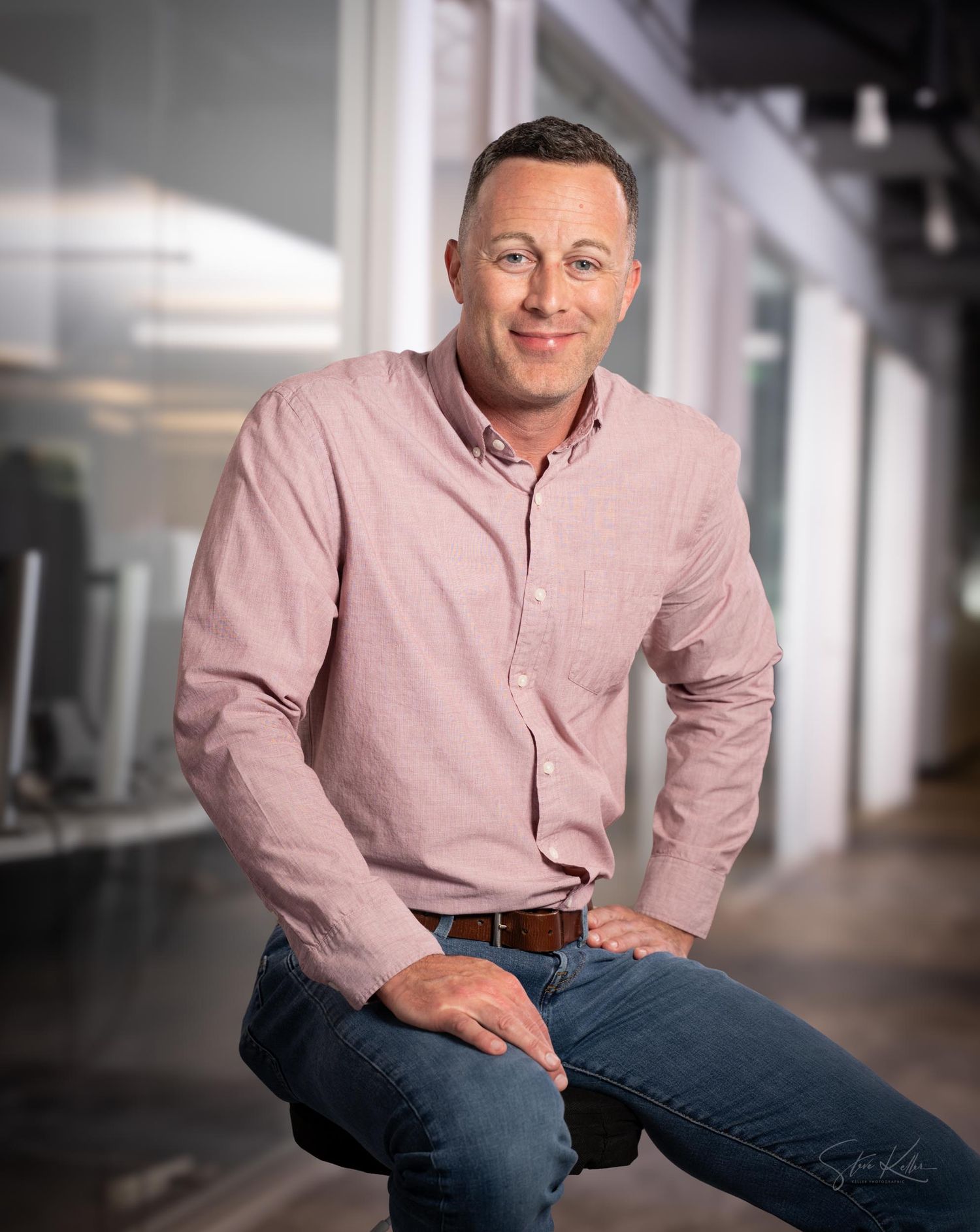 Professional portrait of someone in a pink button-down shirt and jeans seated casually in an office hallway.