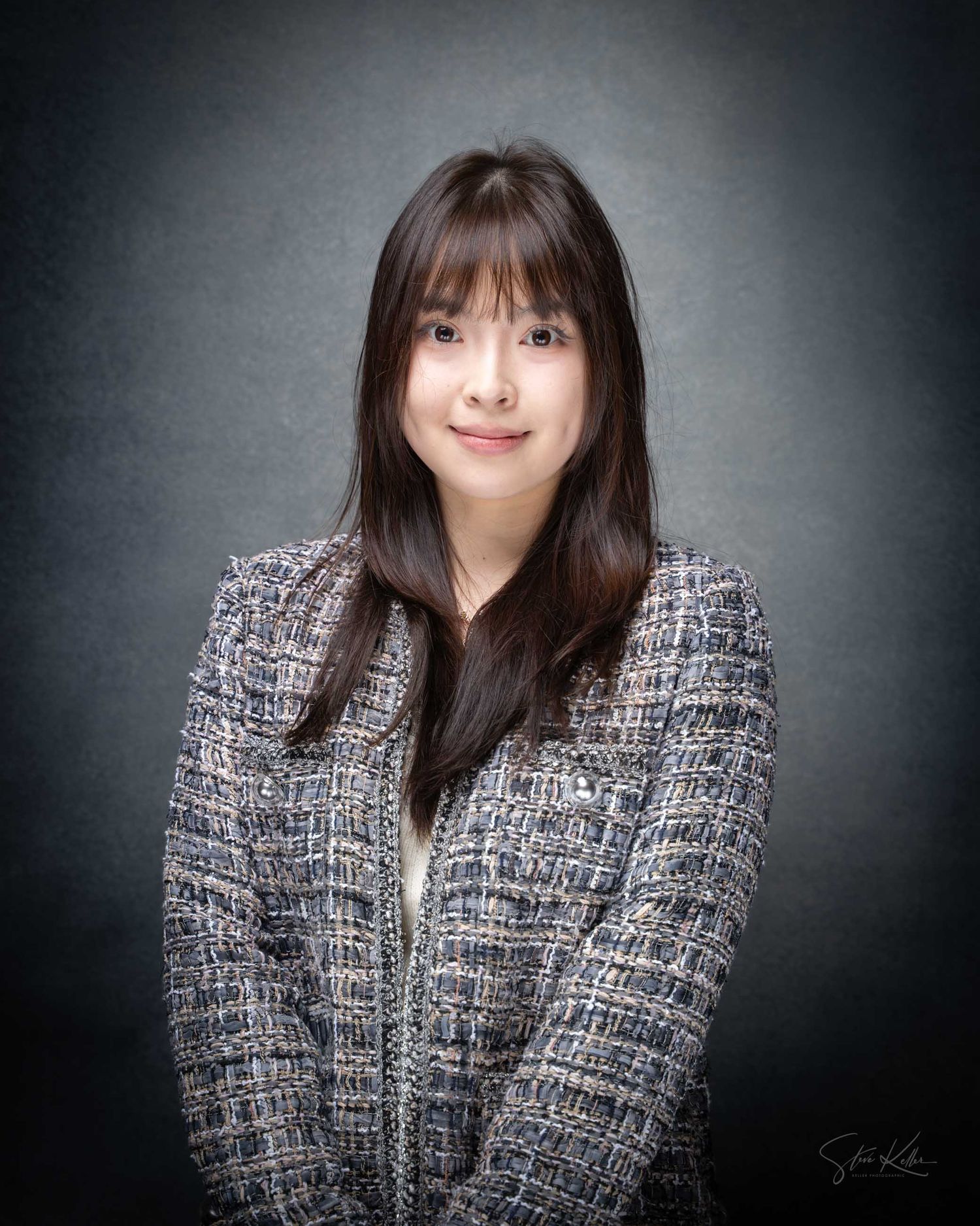 Young Asian woman with dark hair in tweed blazer poses for professional portrait photo against dark background.