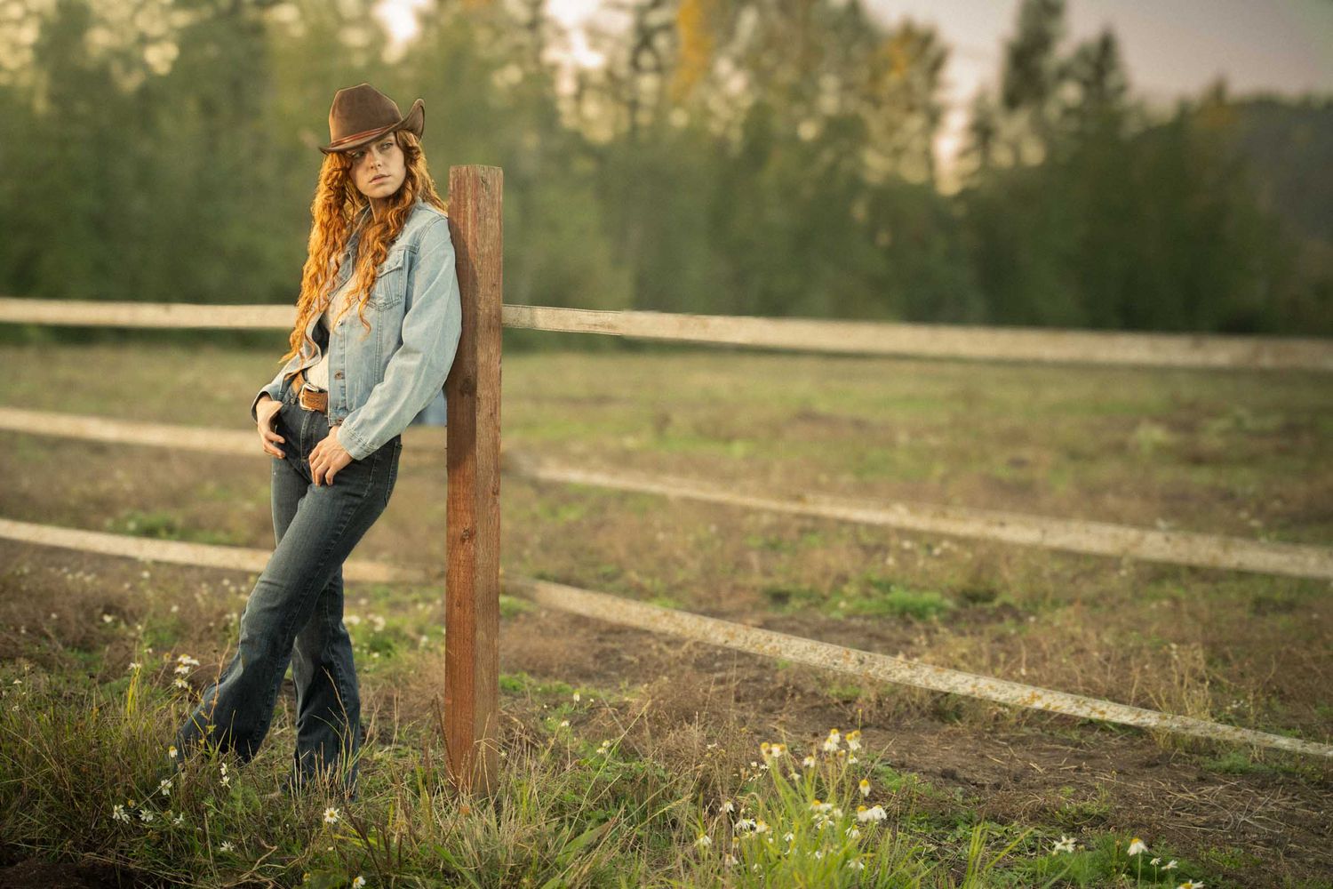 Young woman in jeans and hat leaning against wooden fence post in a rural field at golden hour.
