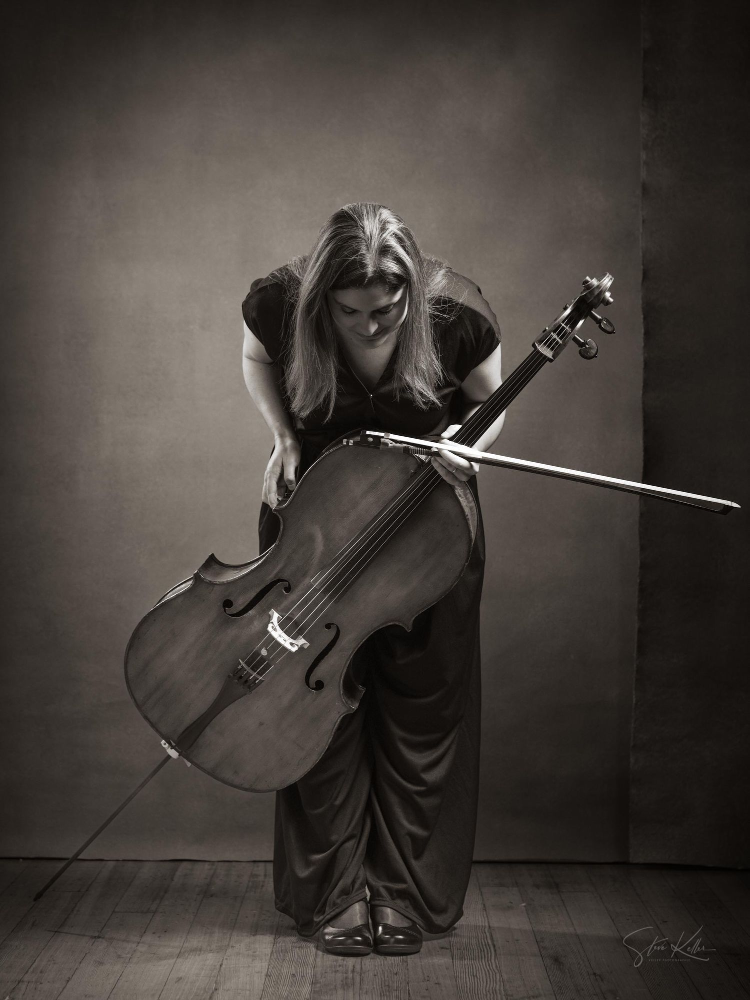 A dramatic black and white photograph of a cellist in a flowing dress bending over their instrument during a performance.