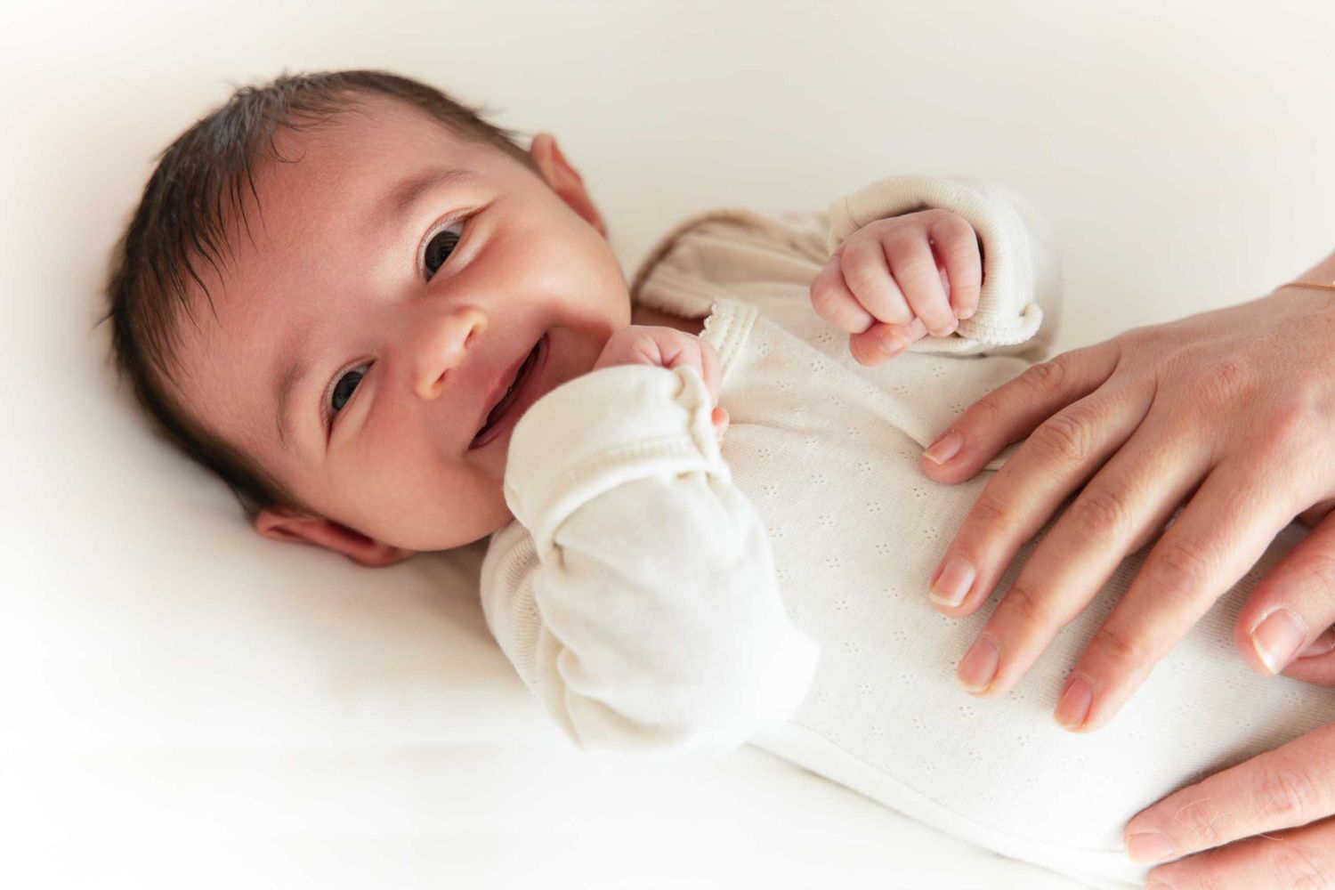 A happy baby in white clothing lies on their back while being held by adult hands.