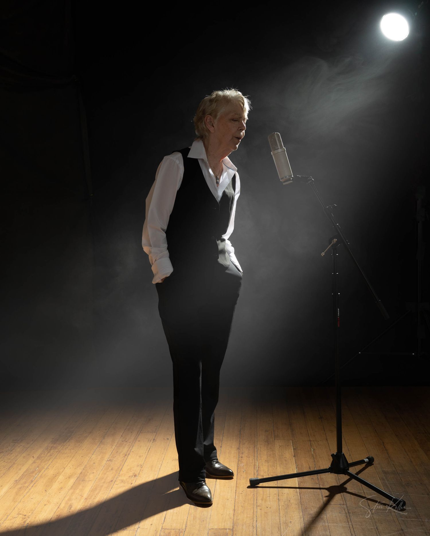 A performer stands in dramatic stage lighting on a wooden floor next to a microphone stand in formal attire.