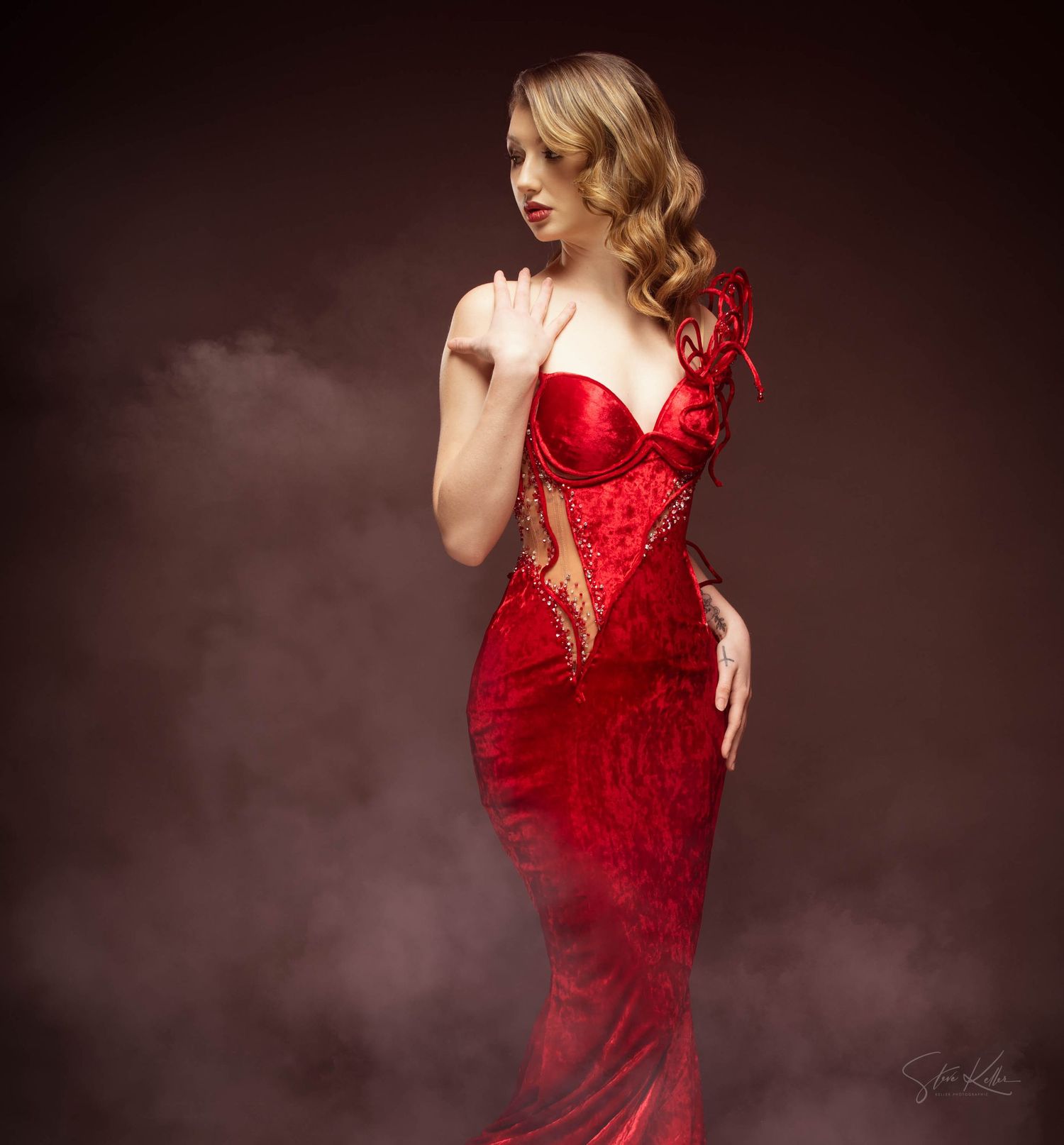 A glamorous figure in a form-fitting red lace gown and blonde wavy hair poses against a dark smoky background.