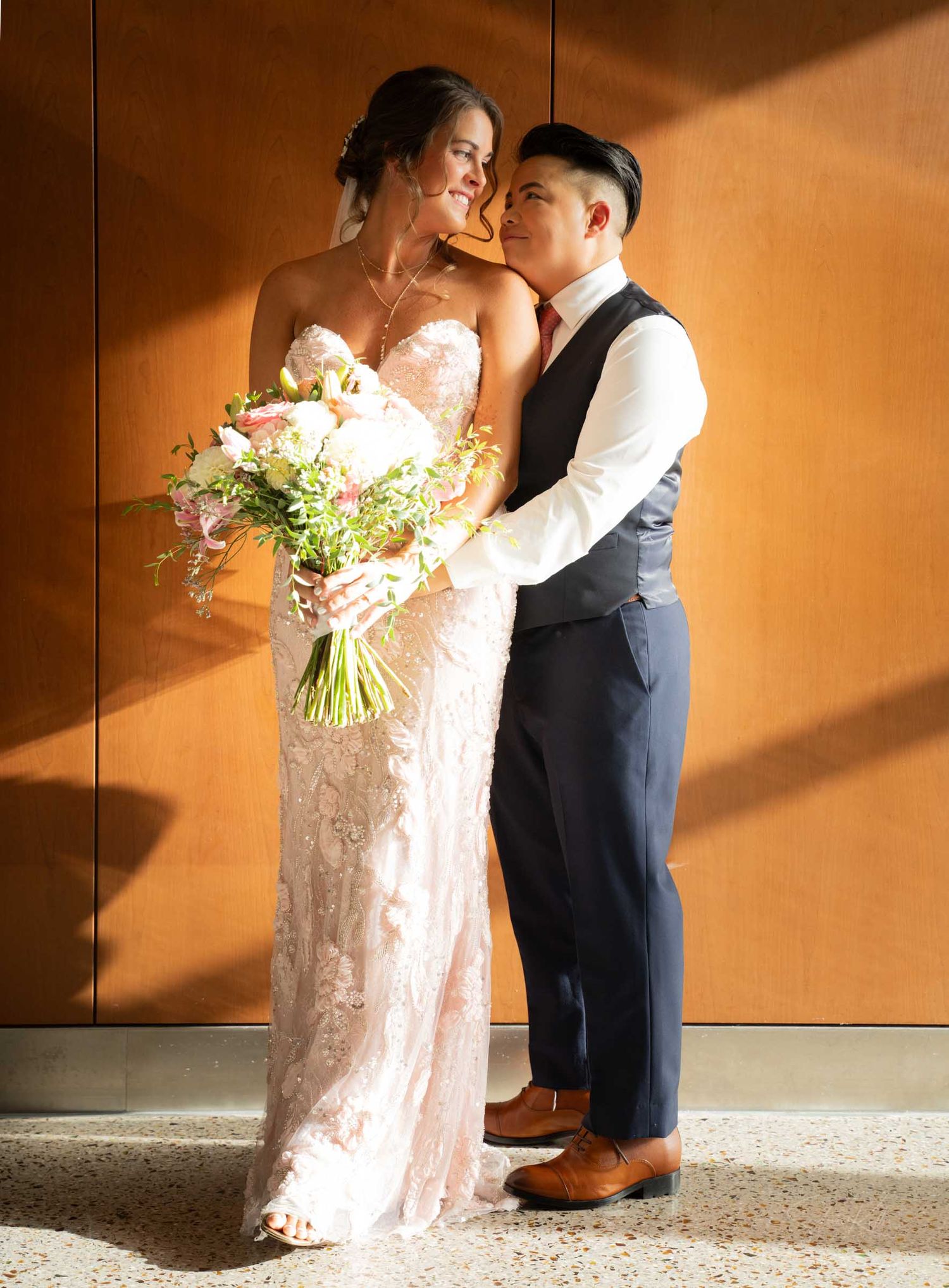 Bride in lace gown holds floral bouquet while groom embraces her against a warm wooden wall.