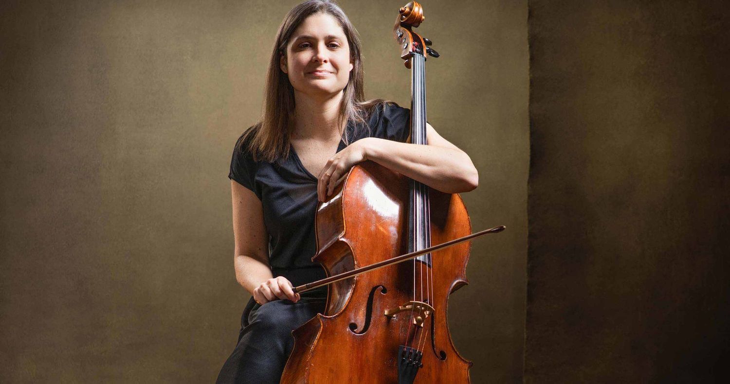 Smiling woman in a black top sits holding a cello and bow in front of a warm, textured golden-brown background.
