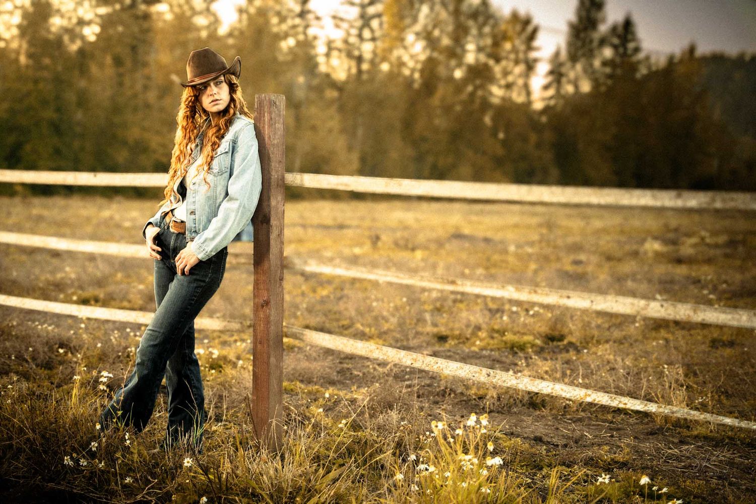 A young woman in a cowboy hat and flare jeans leans against a wooden fence post in a golden sunlit rural field.
