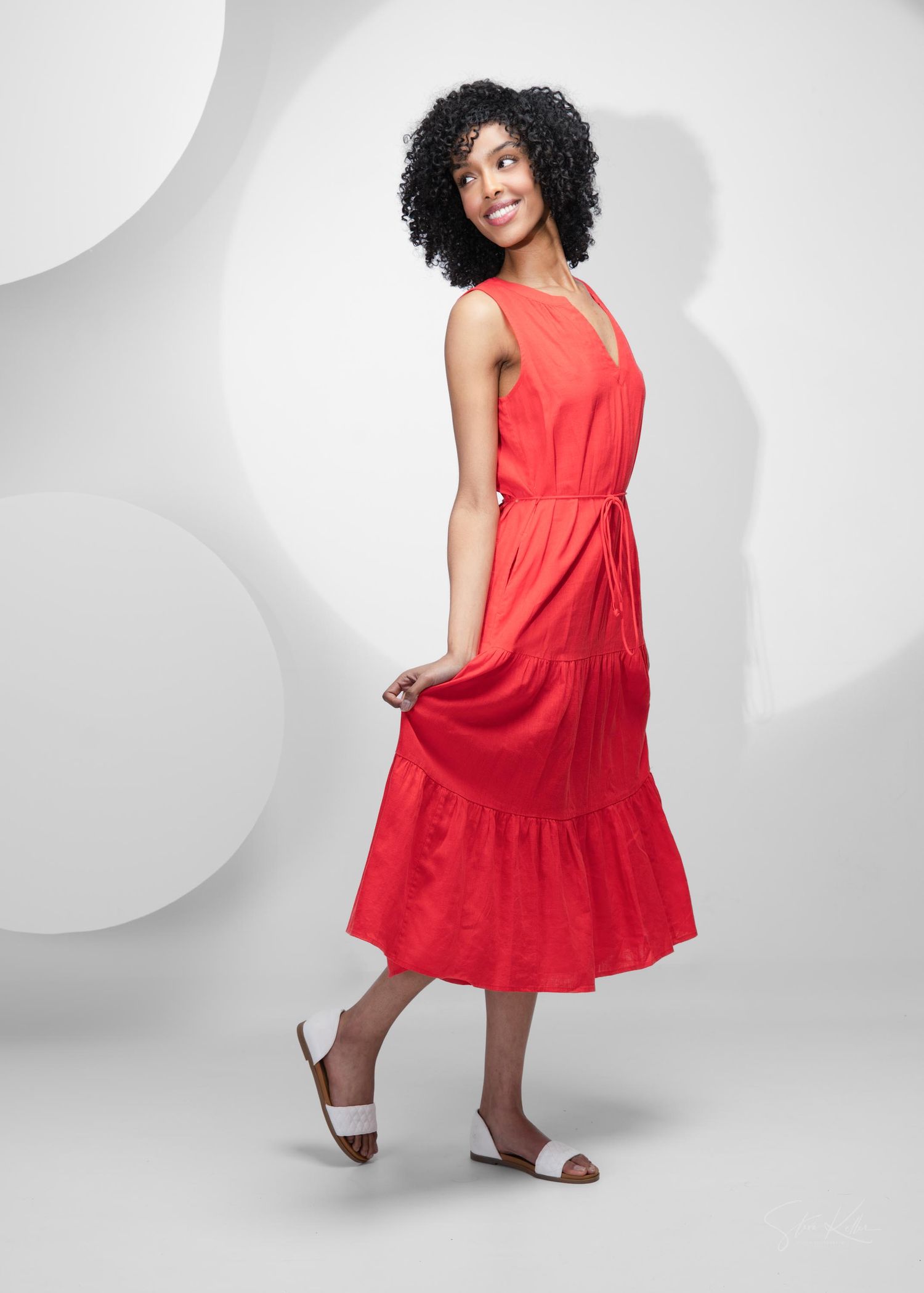 Model wearing a vibrant red sleeveless midi dress with tiered hem and belted waist against a grey background with white circles.