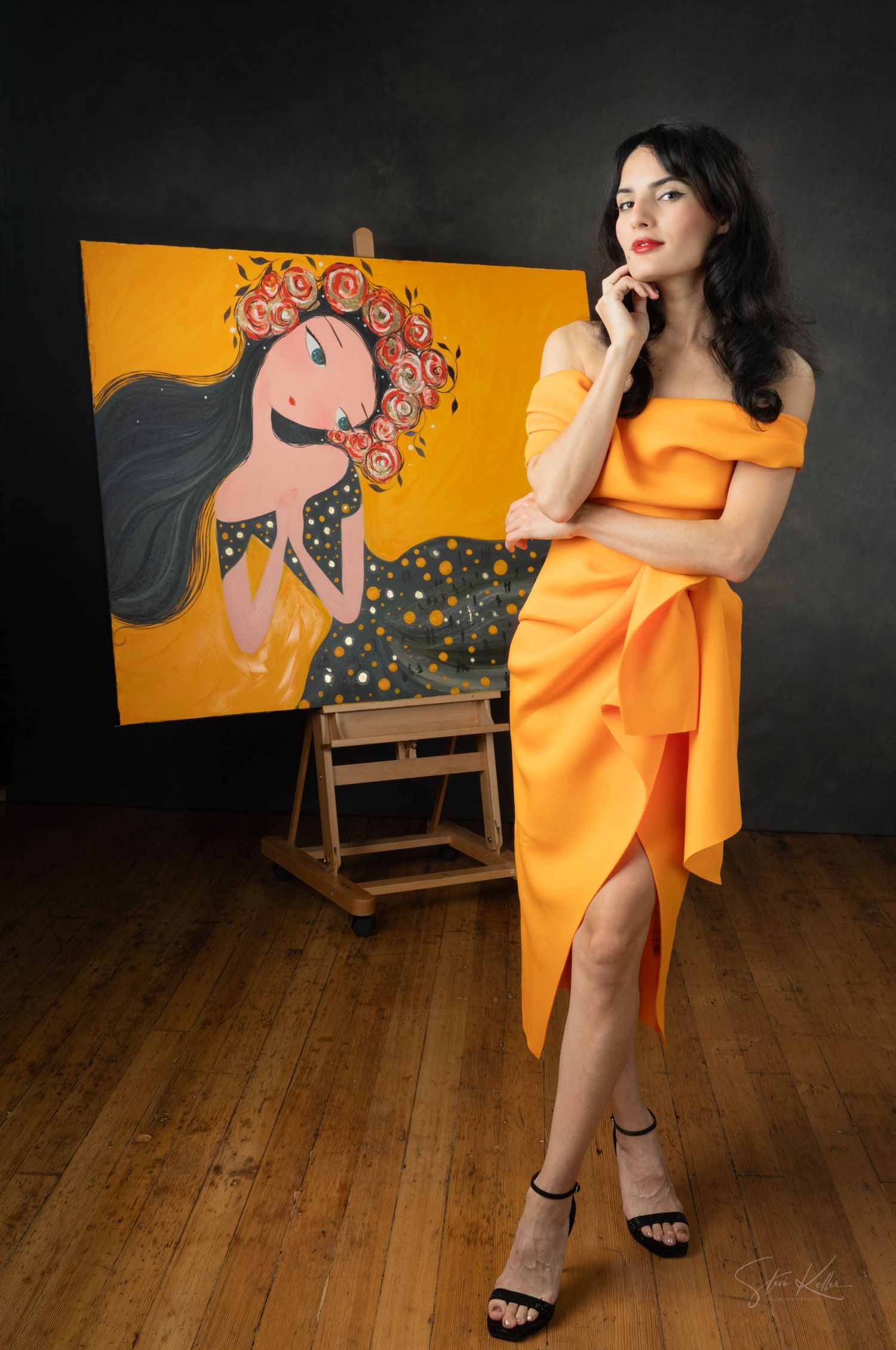 Artist in orange wrap dress poses thoughtfully next to vibrant painting of whimsical figure on yellow background.