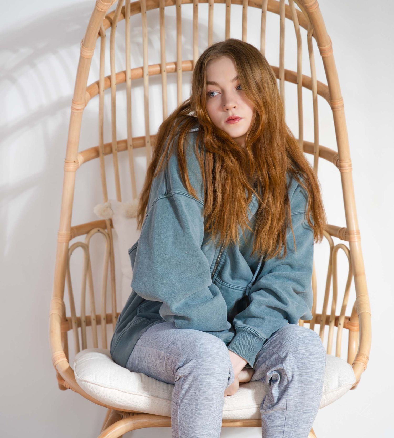 Person in blue sweatshirt and gray pants sitting in rattan hanging chair against white wall.