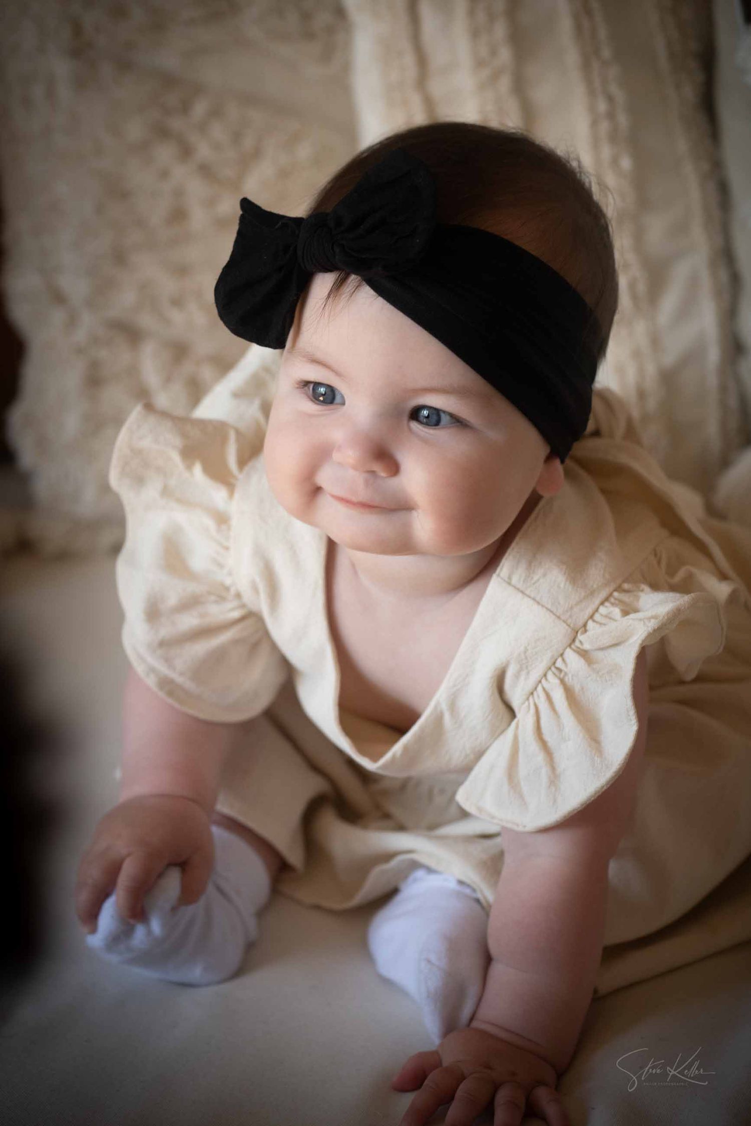A baby in a cream-colored dress and black cap crawls across a light-colored surface.