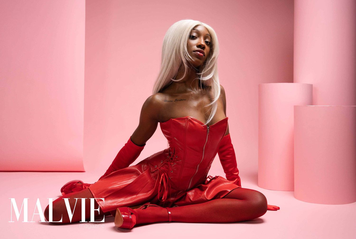 Fashion model in red bodysuit poses elegantly against pink backdrop for Maevie brand photoshoot.
