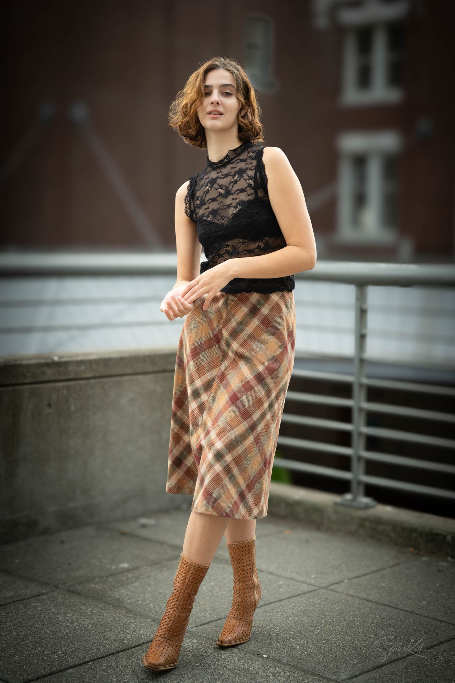 Stylish outfit featuring black lace top paired with plaid midi skirt and brown leather boots on urban backdrop.