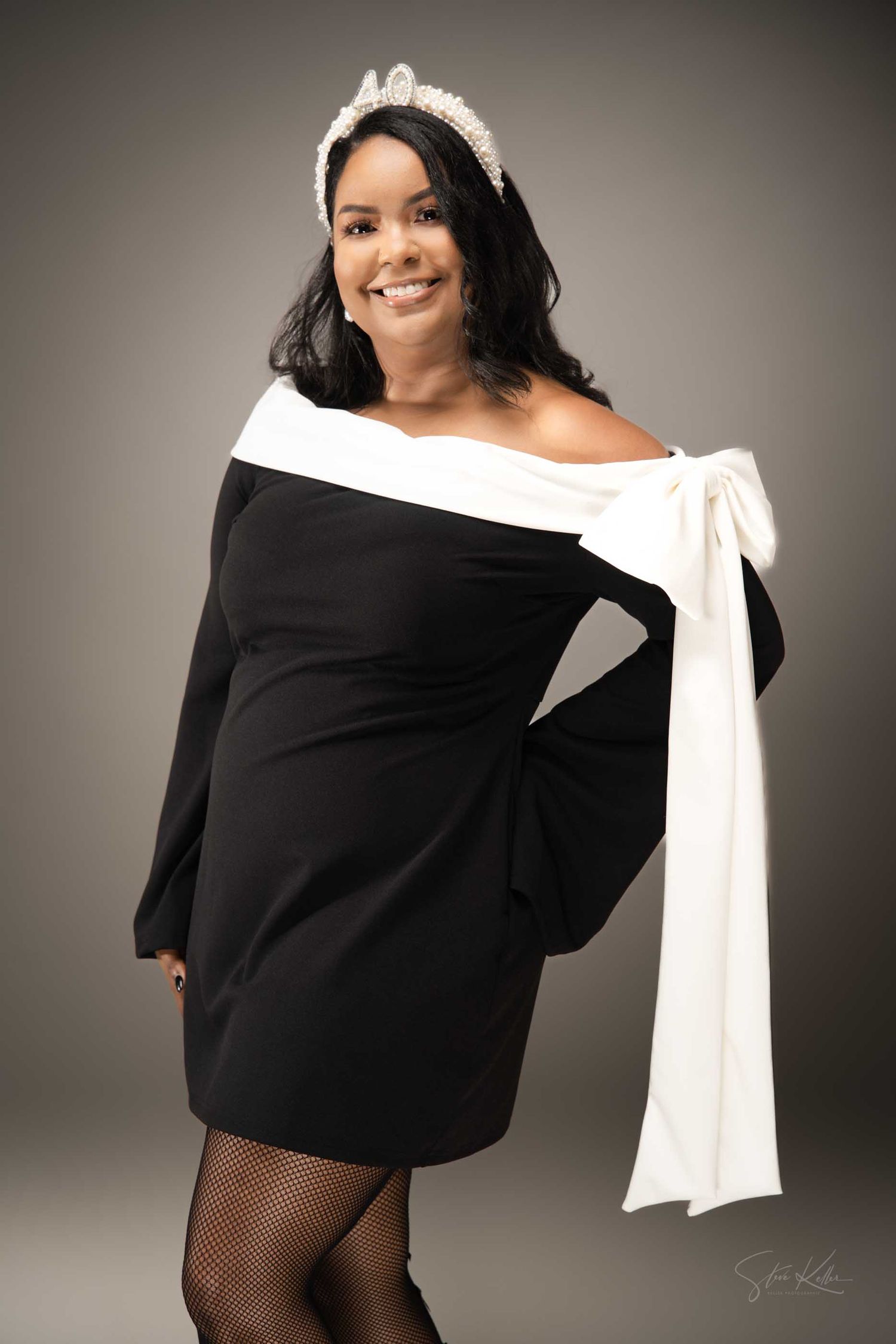 Woman in black off-shoulder dress with white bow and crown accessory, smiling confidently in a studio setting.
