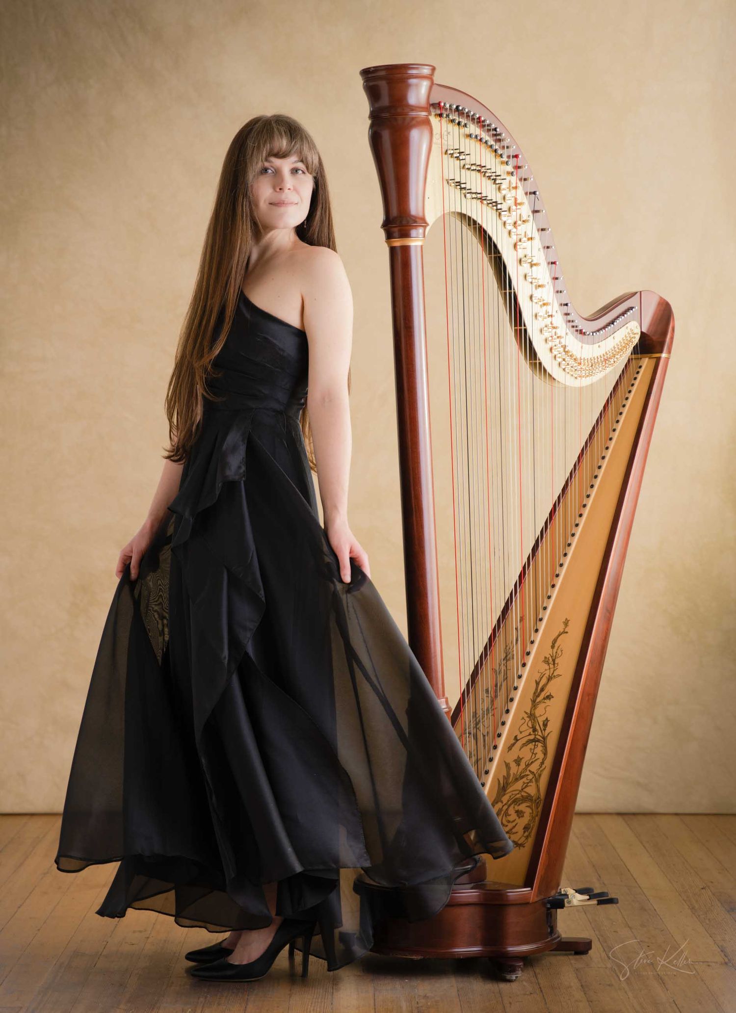 Elegant woman in black strapless gown posing beside a tall wooden concert harp on a warm beige background.