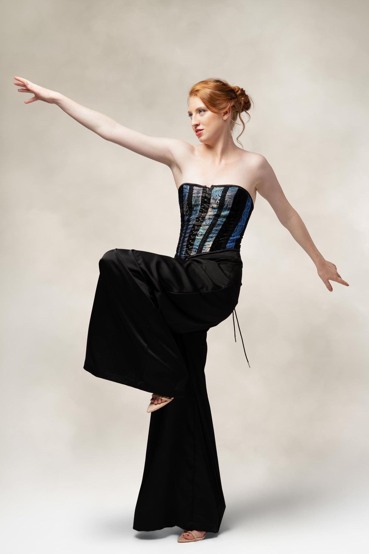 Dancer in black palazzo pants and corset strikes dramatic pose with extended arm against light studio backdrop.