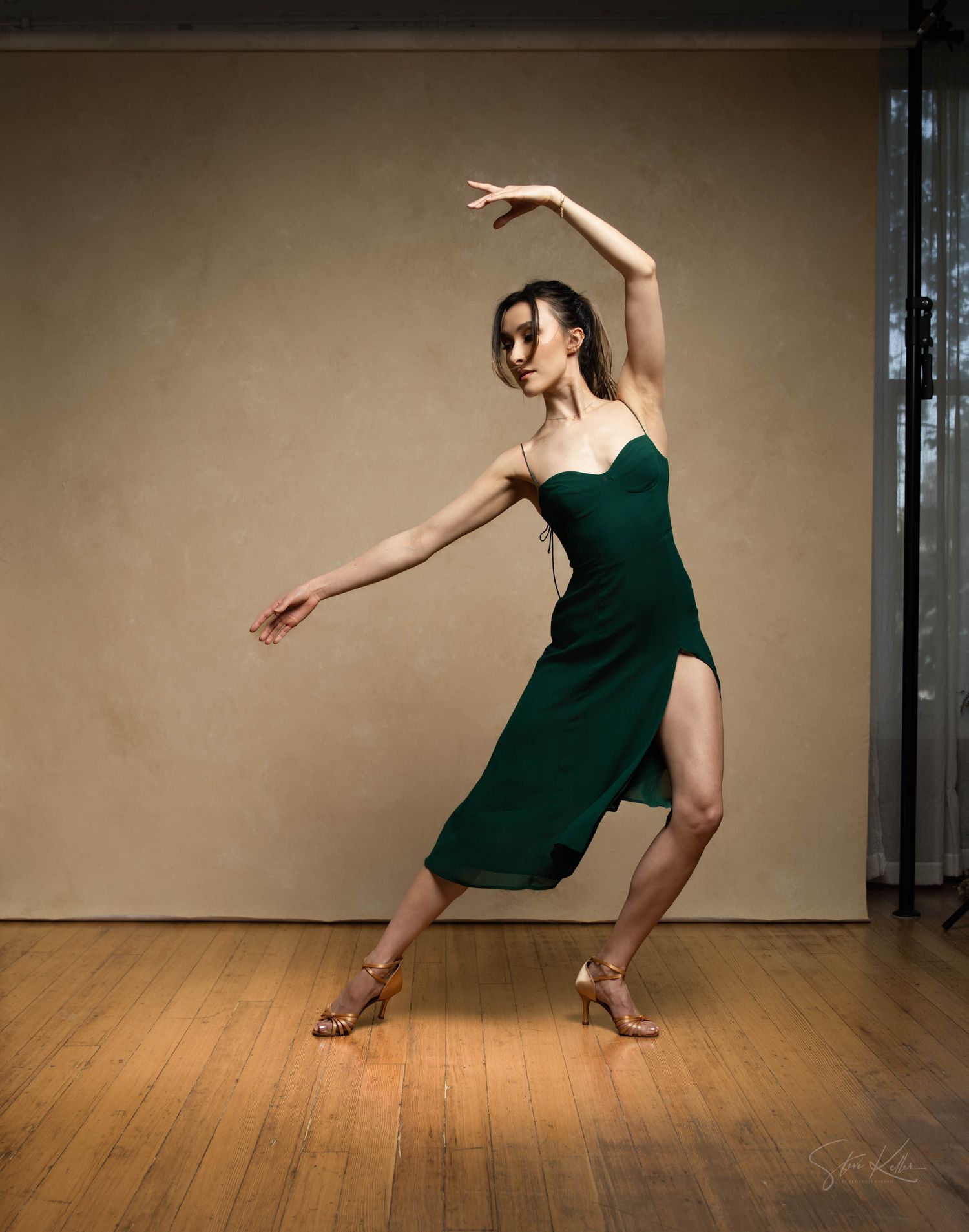 A dancer in an emerald green dress performs an elegant dance pose on a wooden floor against a neutral backdrop.