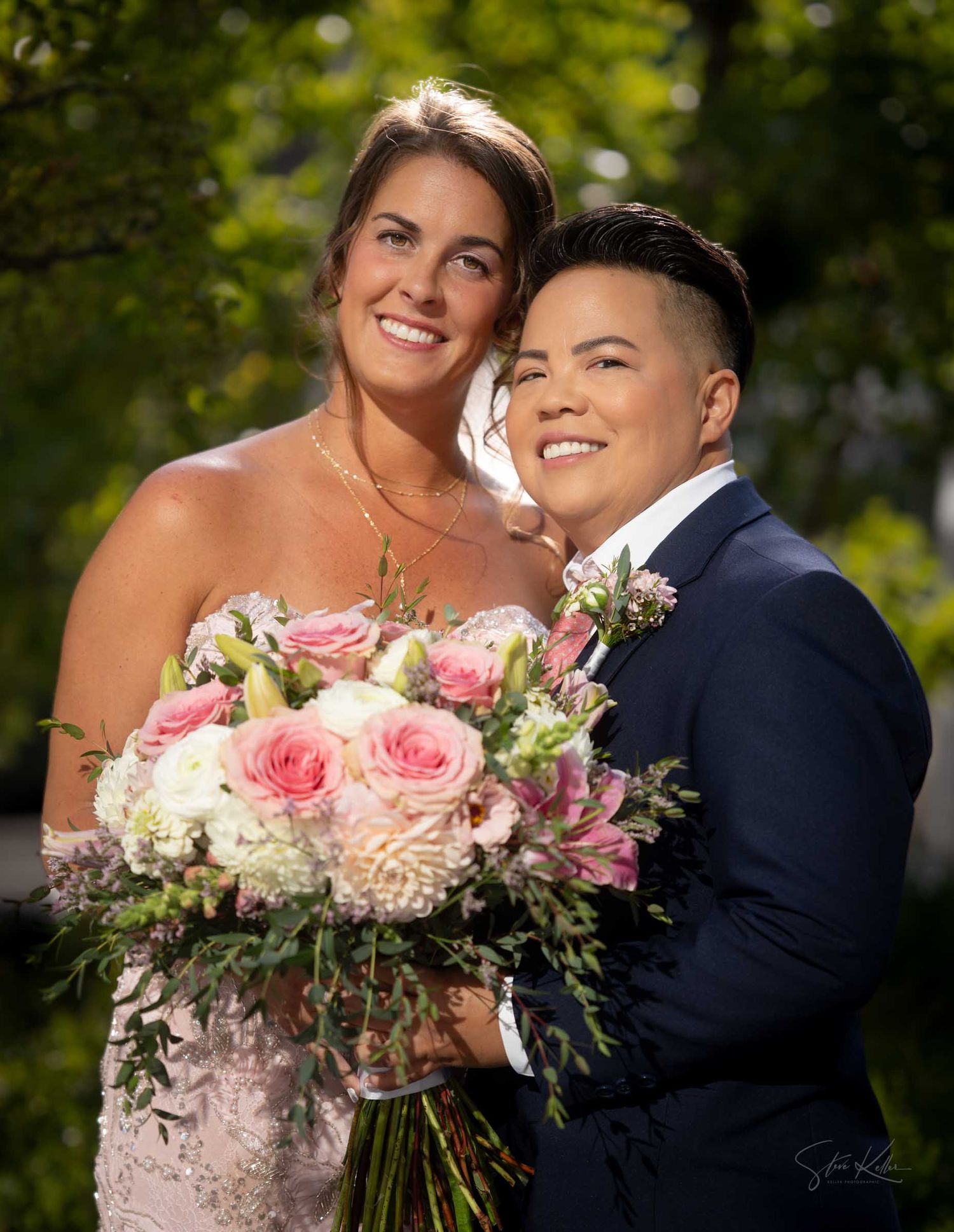 Two brides pose together, one in a pink lace gown holding a bouquet of pink roses and white flowers, the other in a navy suit.